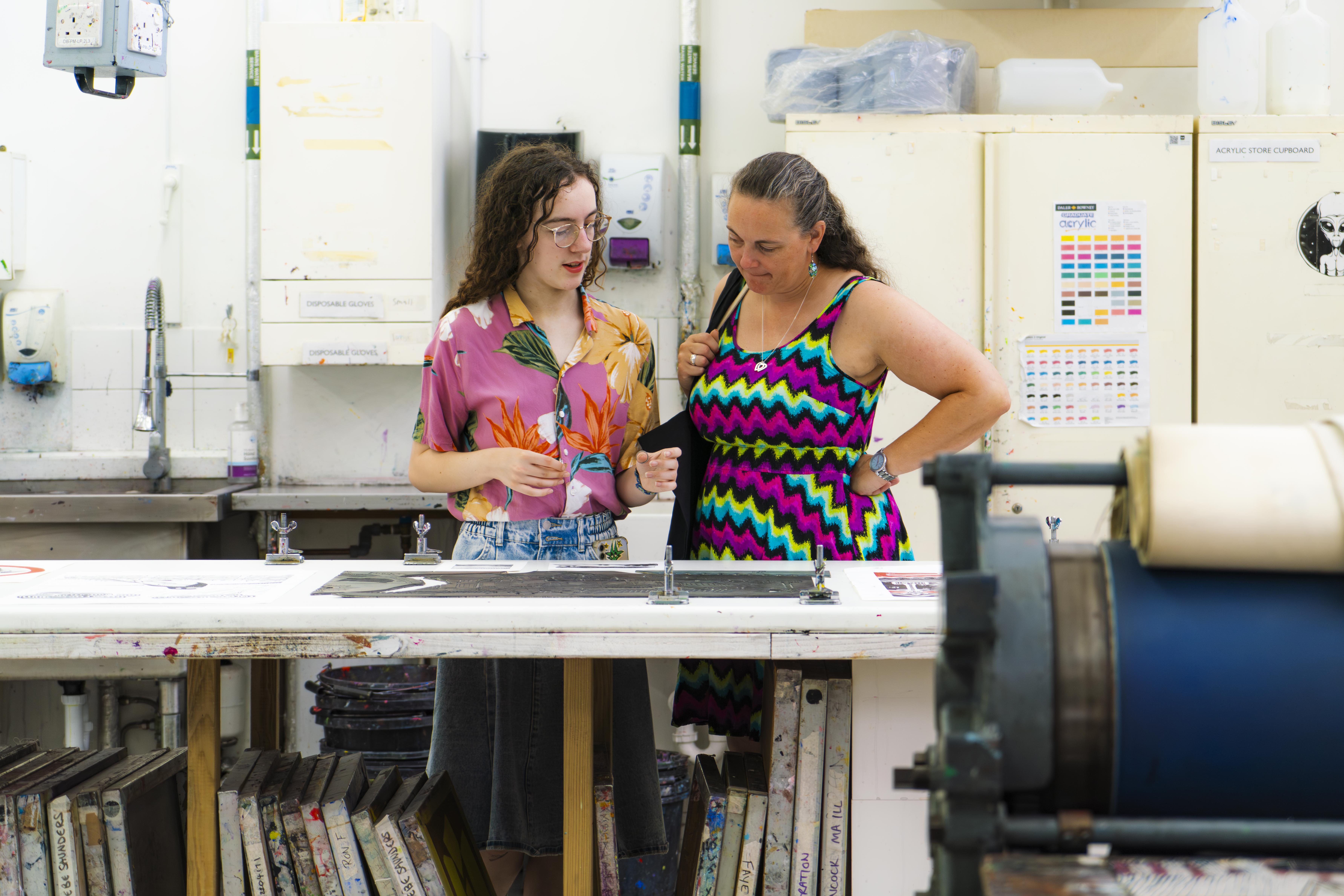 Daughter and mother in bright pink and blue clothing in print studio, looking at print work on a table with canvasses below.