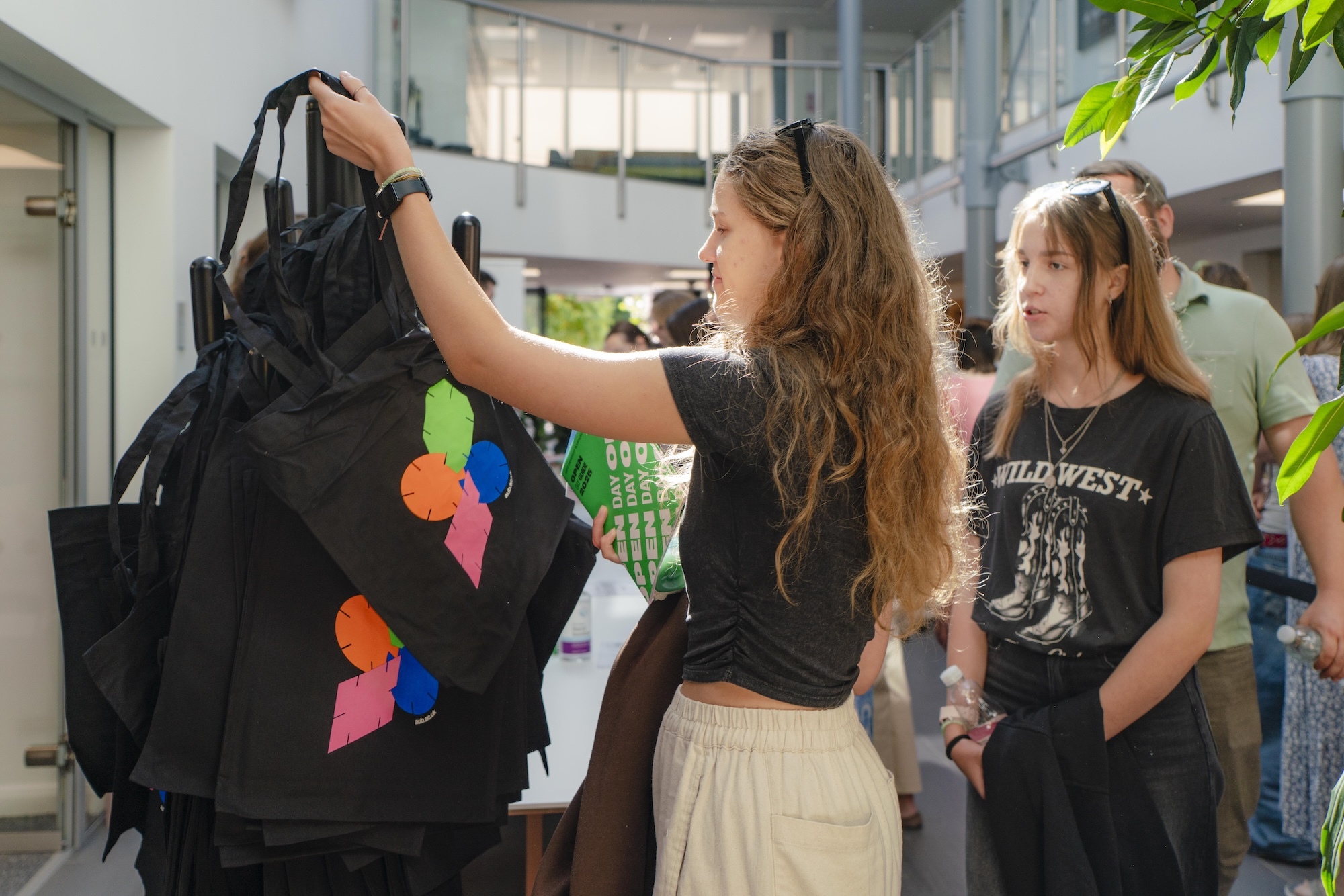 Two young women examining a black garment with colourful geometric shapes in a bright indoor space with glass walls and greenery.