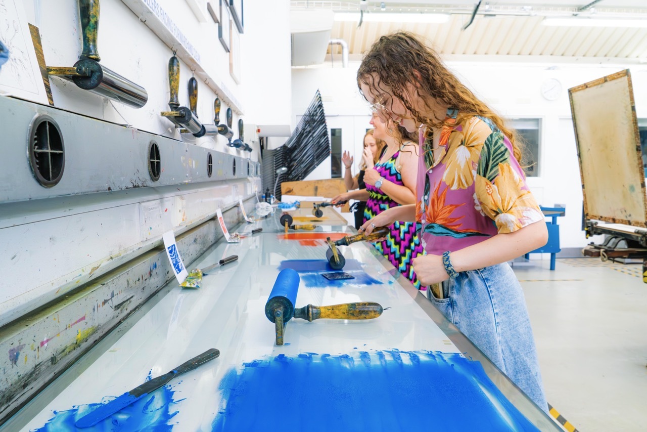 Woman in floral top working on large blue canvas in white art studio with paint tubes and brushes scattered around.