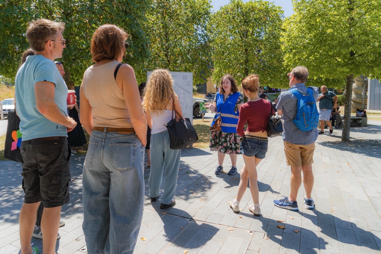 Group of people standing on white stone plaza surrounded by green trees on sunny day, casting shadows on ground.