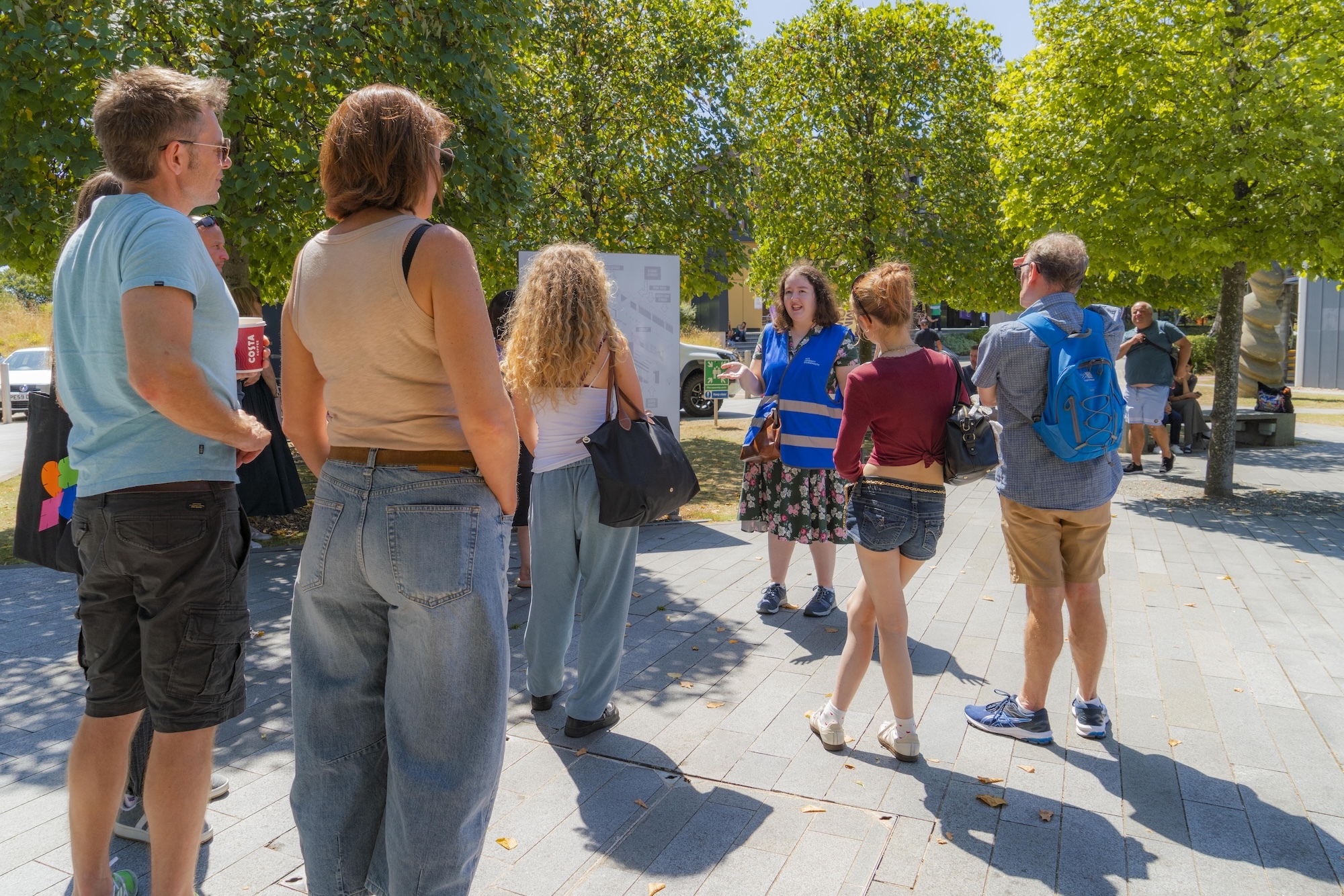 Group of people standing and walking on concrete plaza surrounded by green trees on sunny day.