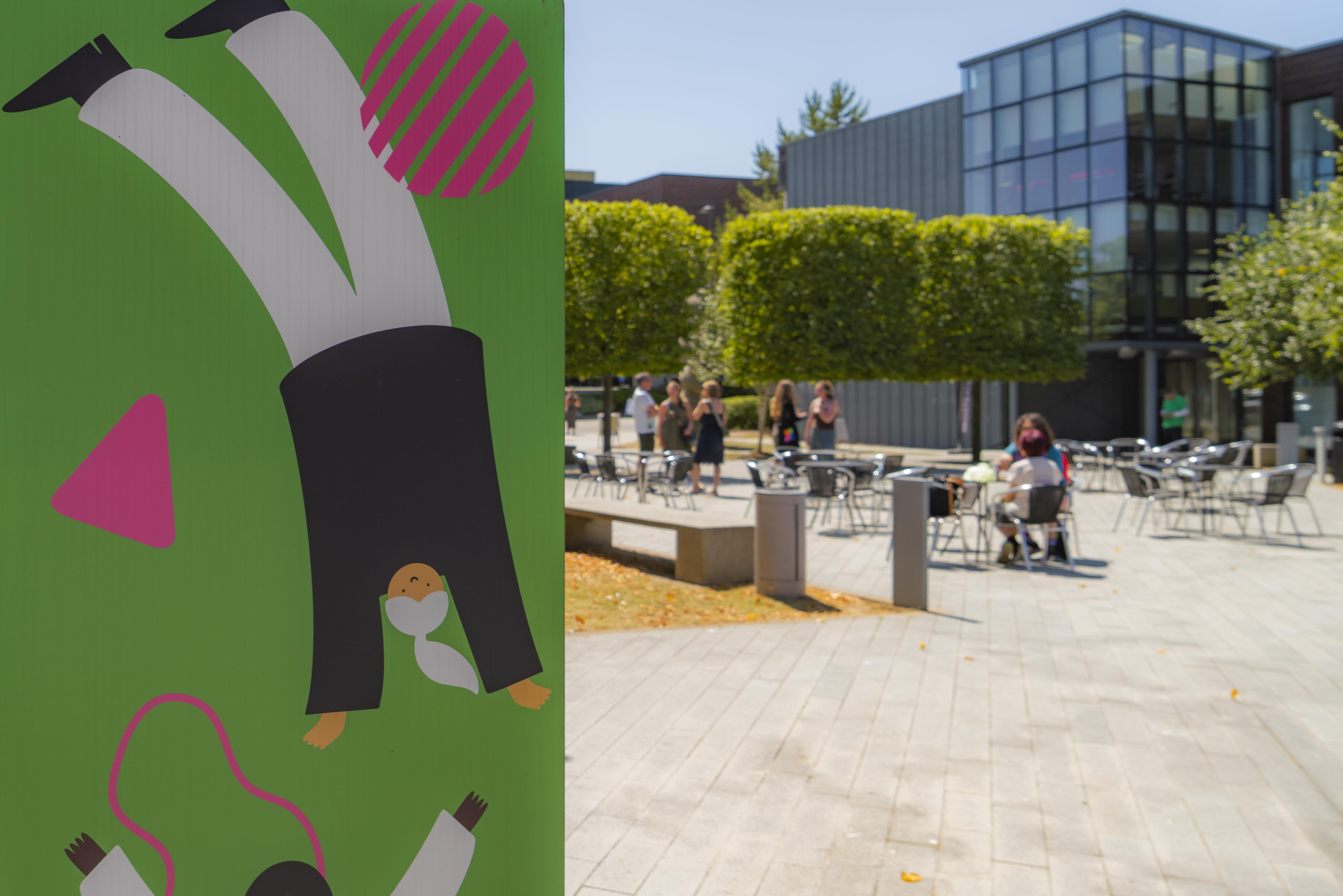 Close-up of a wall panel with green background with pink circles, and an illustration of a person dressed in black and white doing a handstand; with a courtyard part of Arts University Bournemouth campus in the background