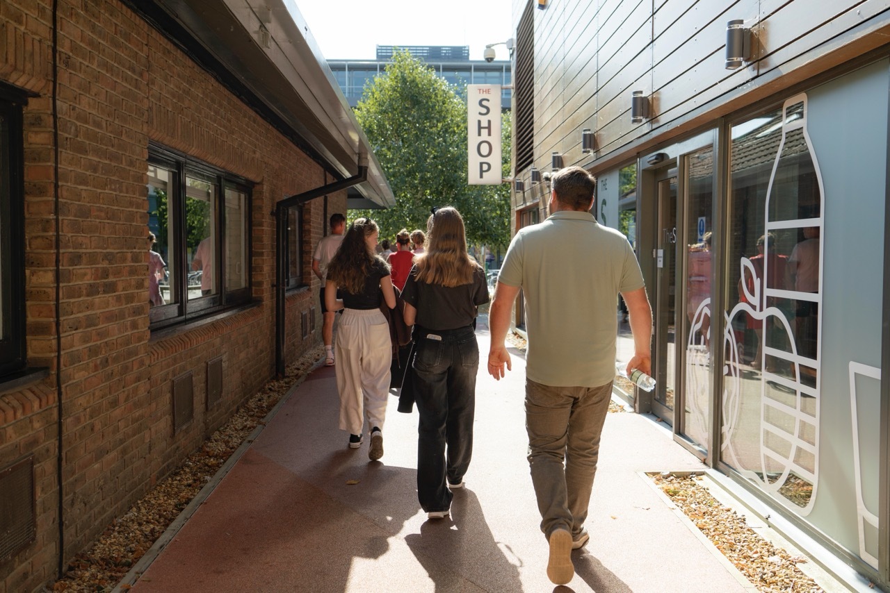 People walking through narrow outdoor shopping corridor between brick and light blue buildings with overhead power lines and shop sign.