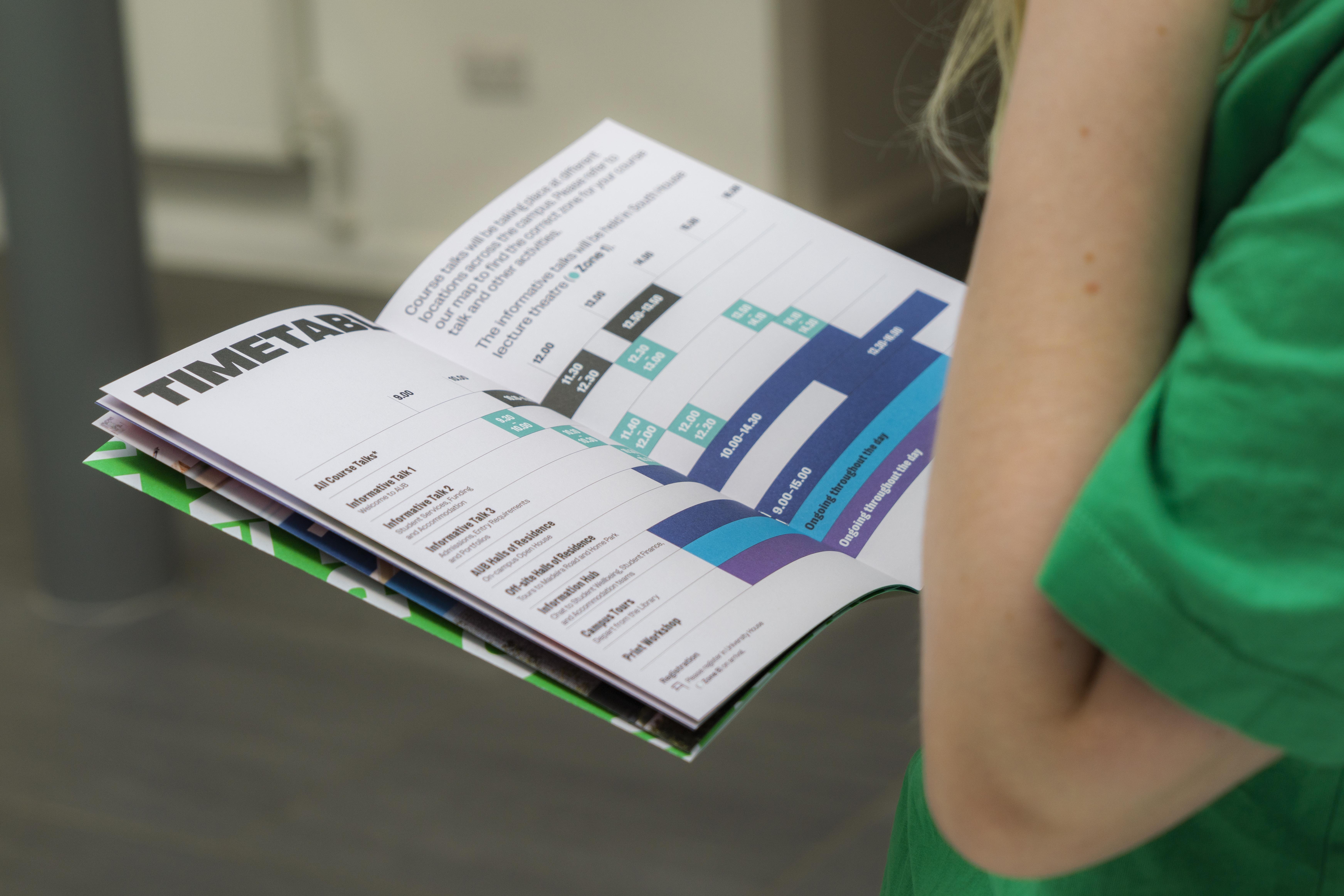 Person in green top holding open timetable booklet showing blue and green bar charts and schedule grids on white pages.