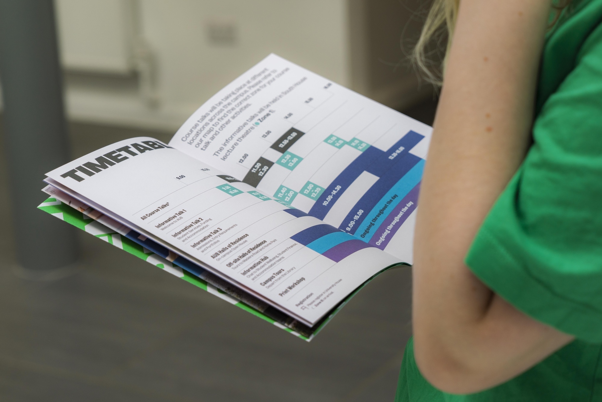 Person in green top holding open magazine showing pages with blue and teal bar charts and "TIMETABLE" header text.