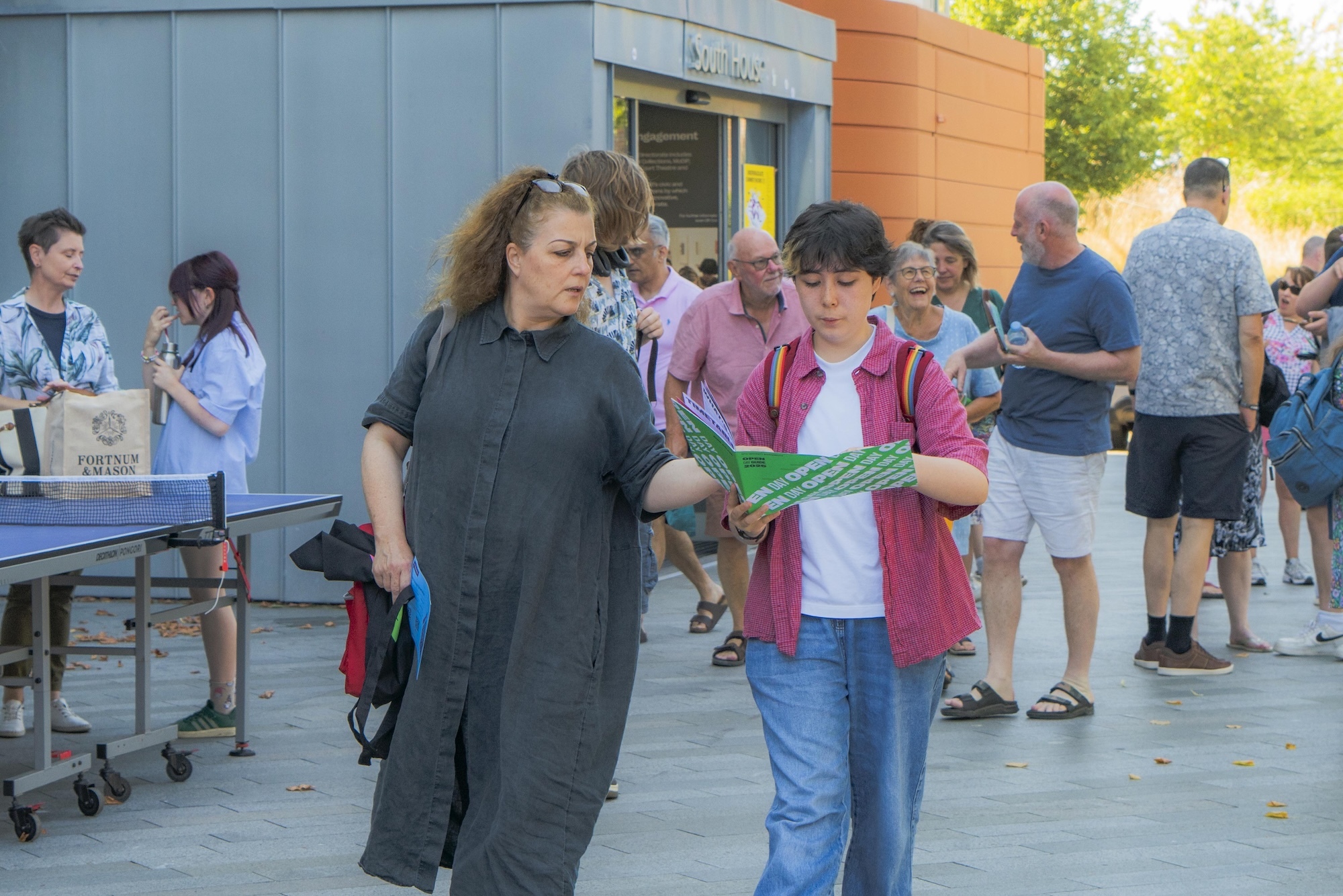 Two people walking; the person on the right is holding green folder outside modern university building with crowd gathered.