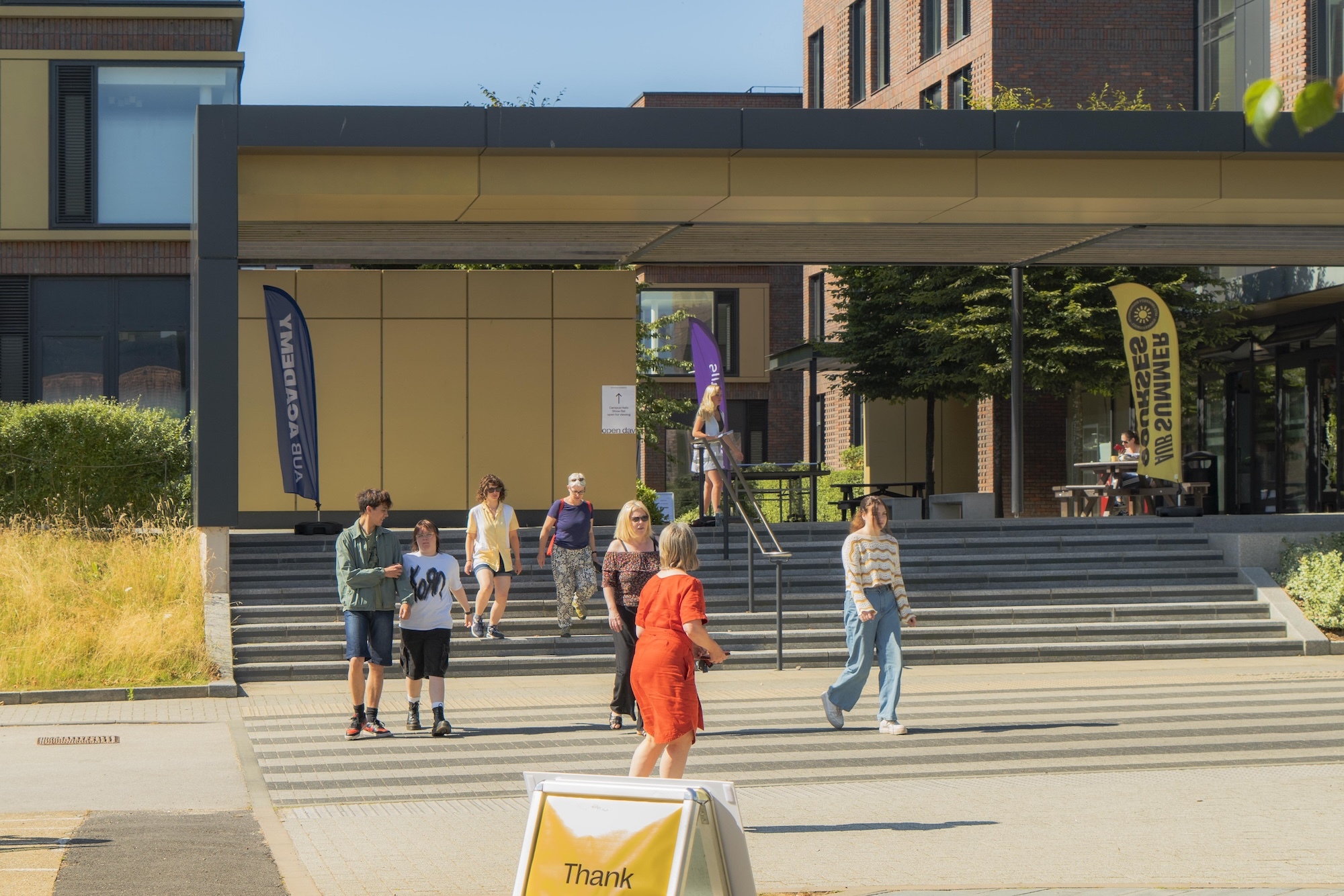 Children and adults on concrete steps and plaza area in front of modern building with glass and brown panels under blue sky.