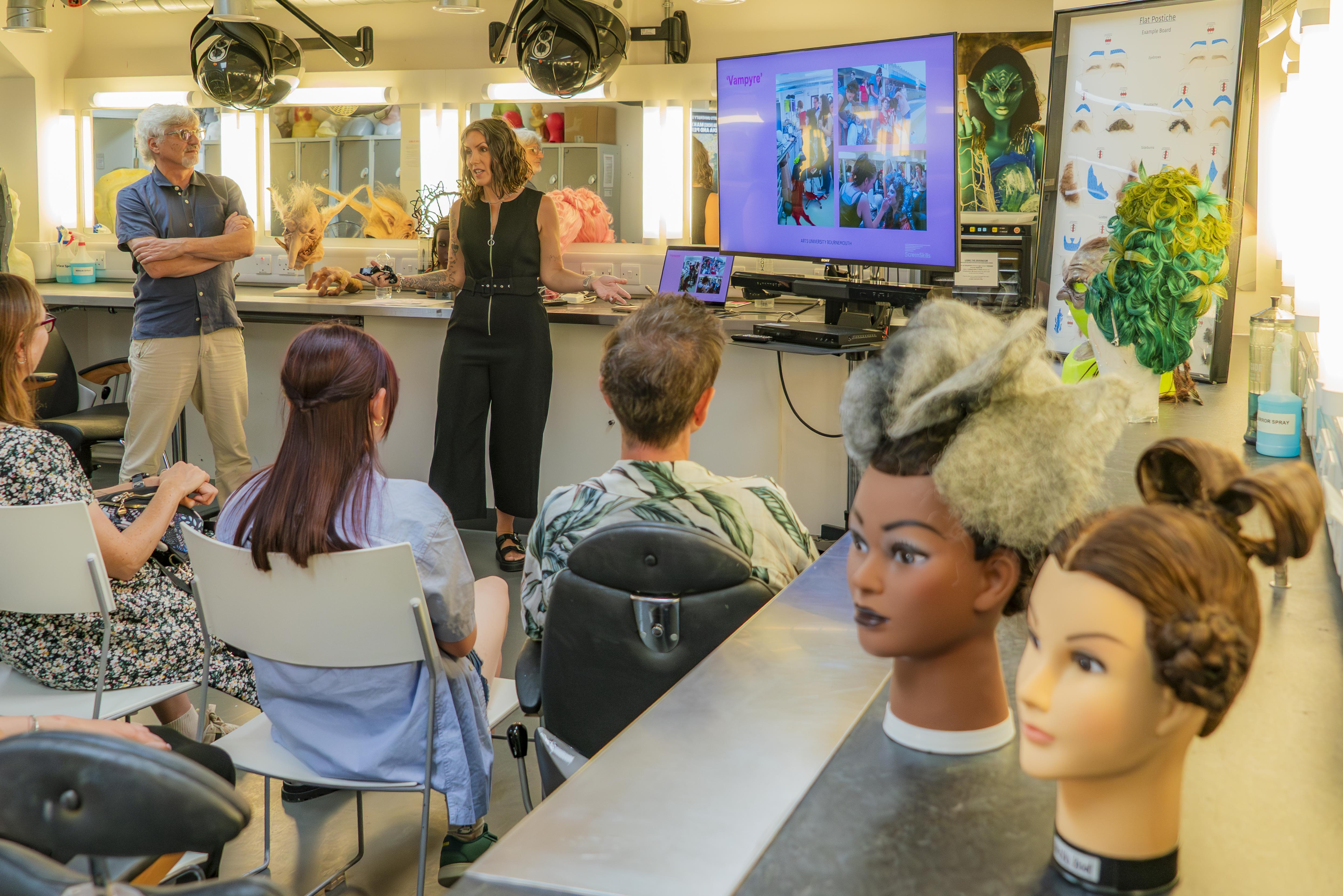 BA (Hons) Make-up, Hair and Prosthetics classroom with visitors sitting and listening to a talk, mannequin heads in foreground, wall-mounted screen
