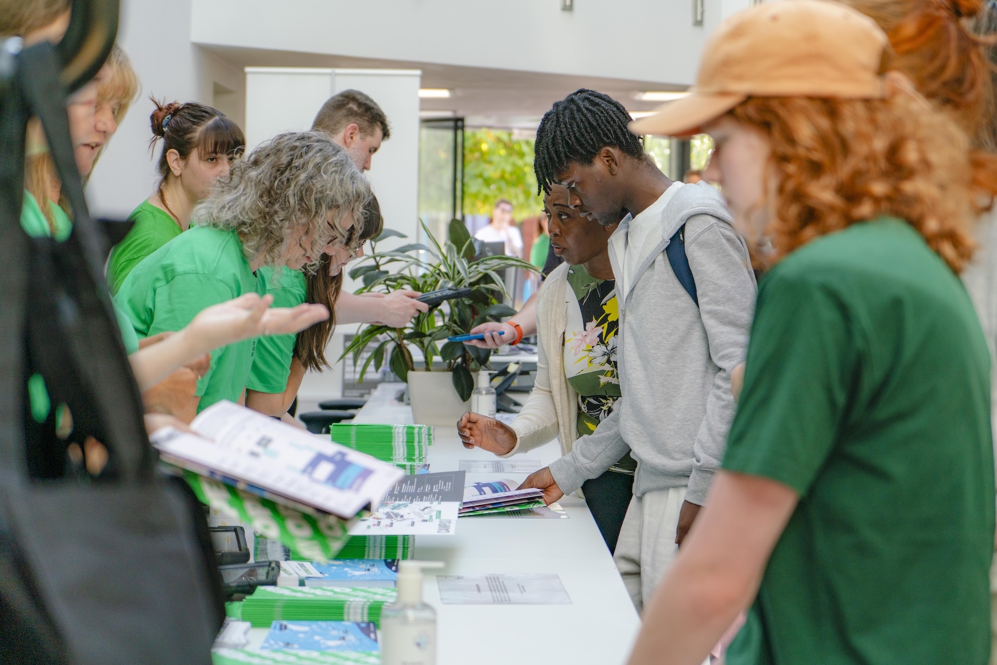 Group of people in green shirts gathered around white tables with papers and materials in bright indoor space with windows.