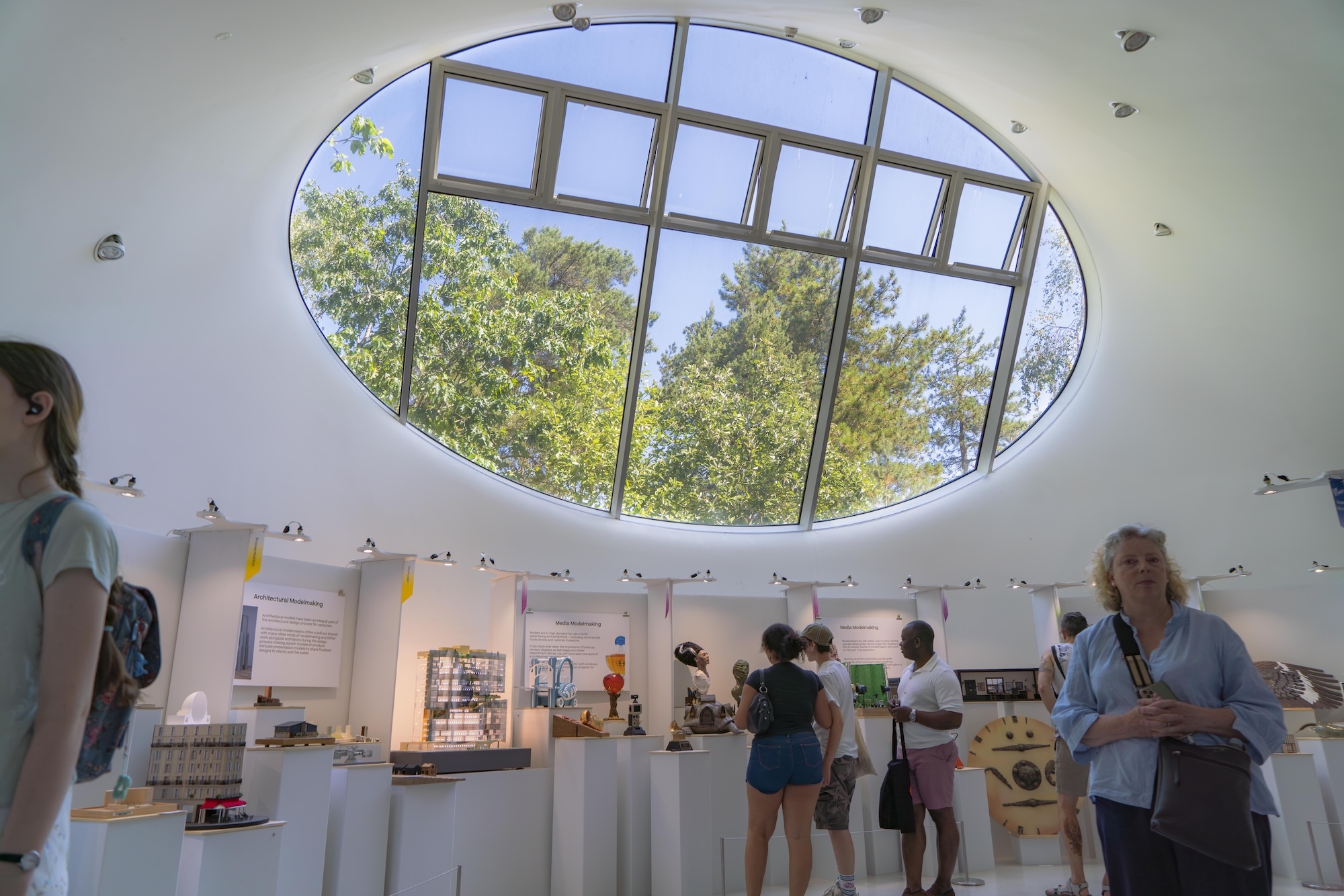 White interior space with large oval skylight showing blue sky and green trees. People browsing displays below curved white ceiling.