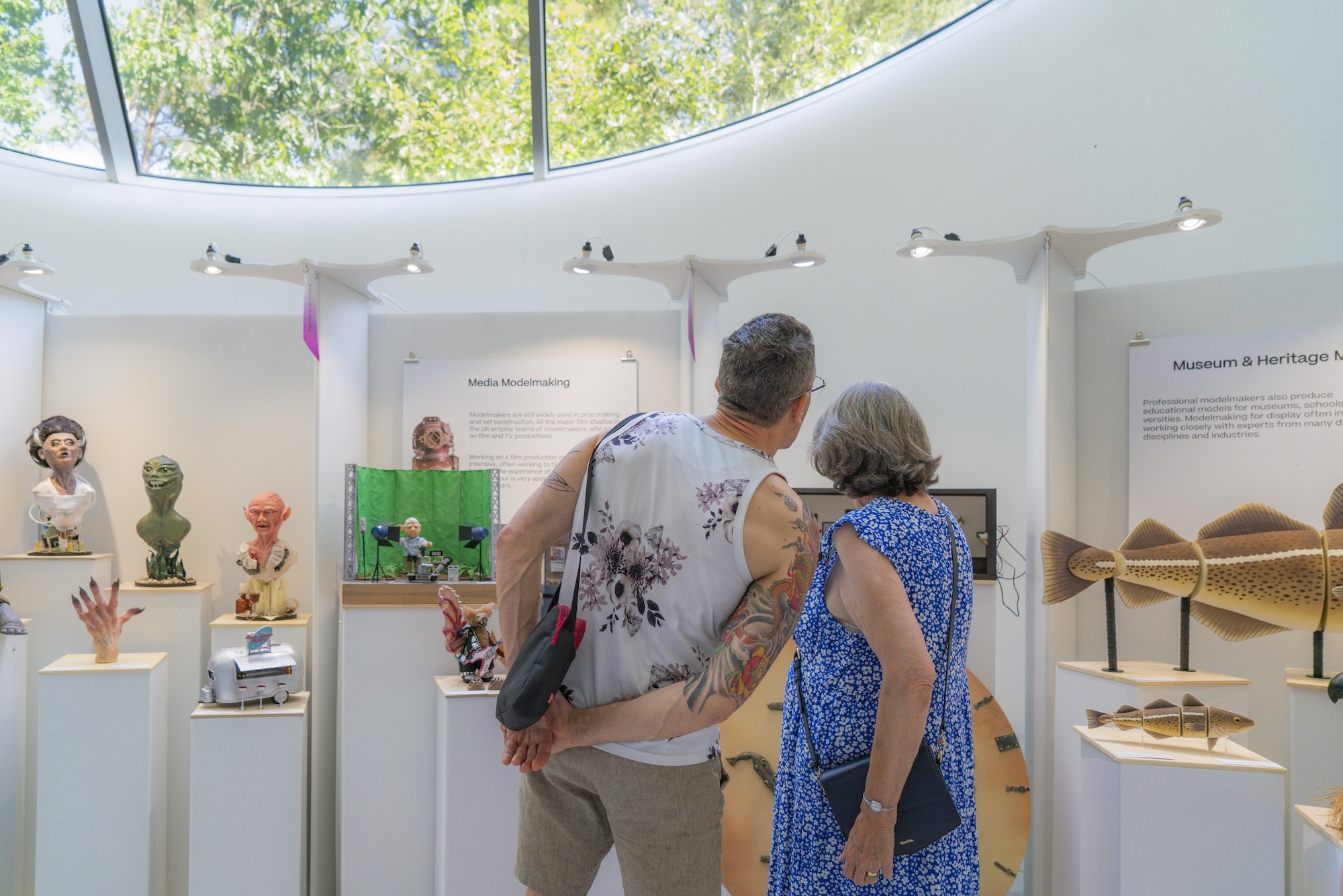 Two visitors viewing display at AUB Summer Shows with colourful sculptures and artefacts in white curved gallery space with skylights.