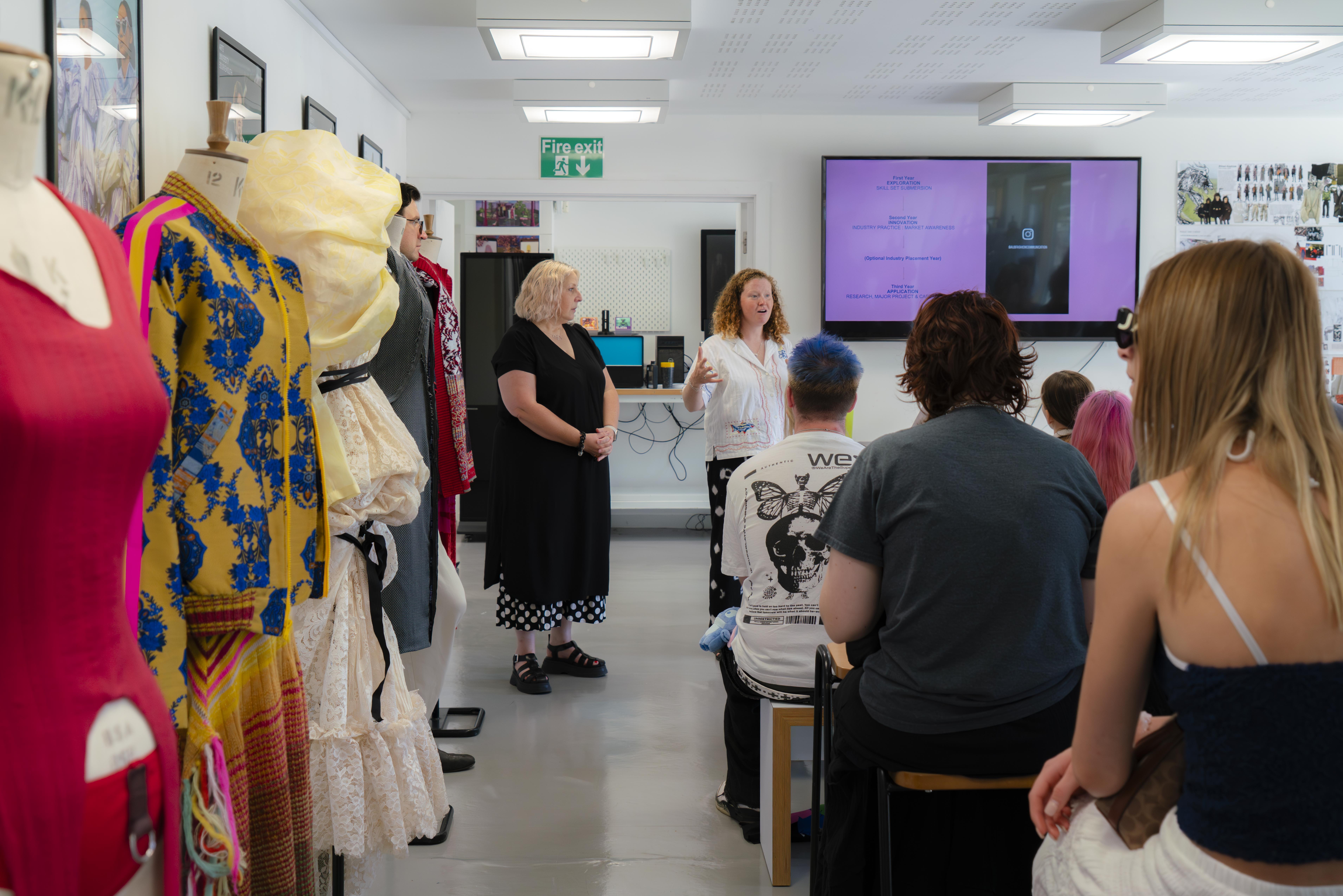 Classroom with clothing display rack on left, E Crehan and C Budnarowska presenting to seated Open Day visitors, purple screen on wall, fluorescent lighting.