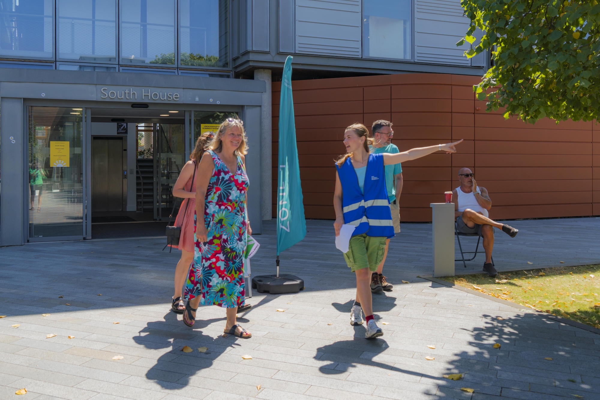 Two people walking past modern building with glass frontage and red brick section. Man in high-vis vest pointing, seated person visible.
