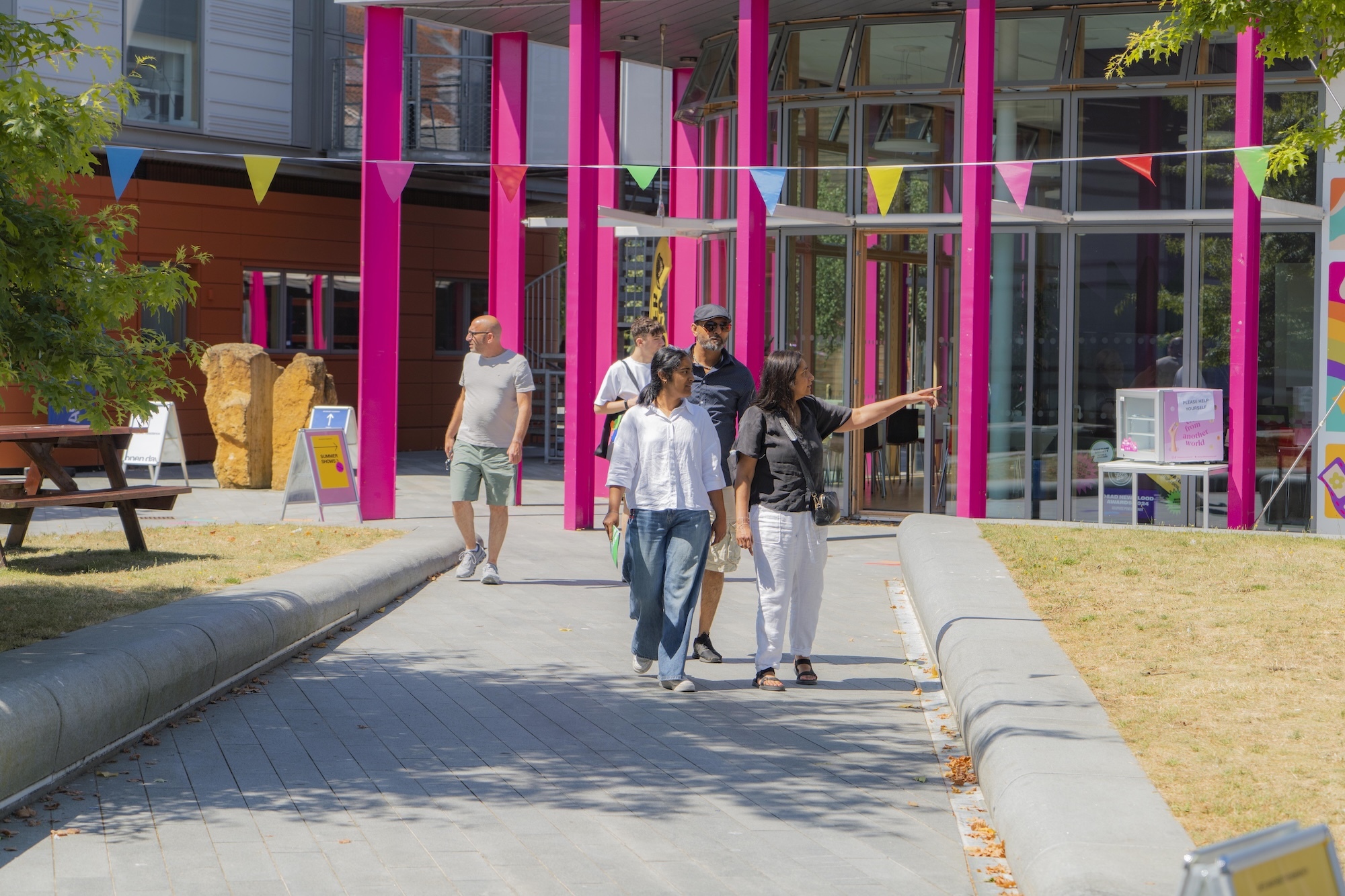 Modern building with bright pink columns and glass facade. People walking on concrete path with colourful bunting overhead.