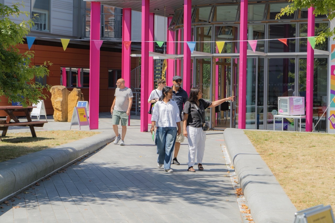 Modern building with bright pink vertical columns and glass facade. People walking on concrete path with colourful bunting overhead.