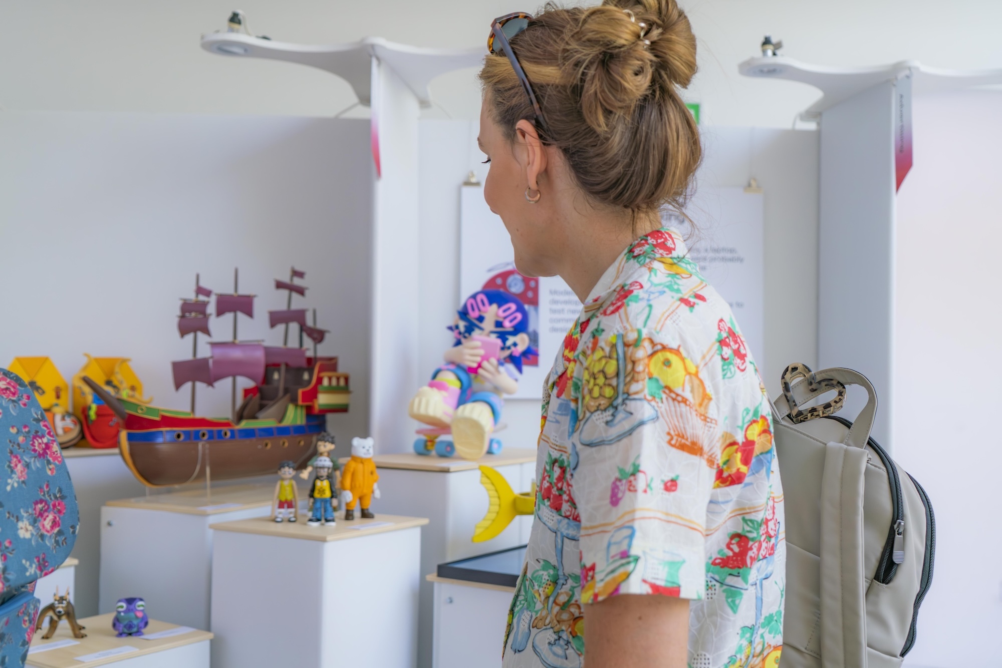 Woman with blonde hair bun wearing colourful floral shirt stands at white shelving unit displaying various toys and decorative items.