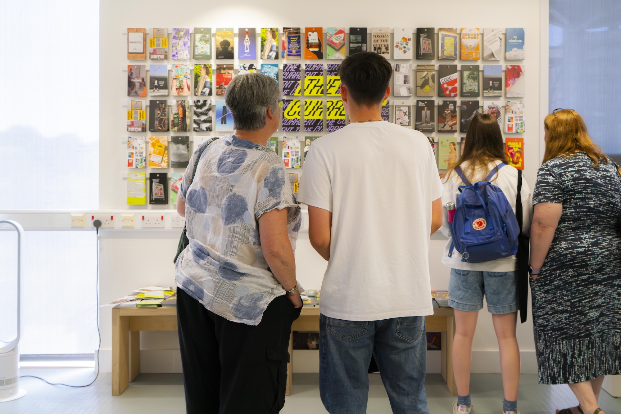 Four people viewing wall display of colourful book covers and posters in gallery space with white walls and wooden bench.