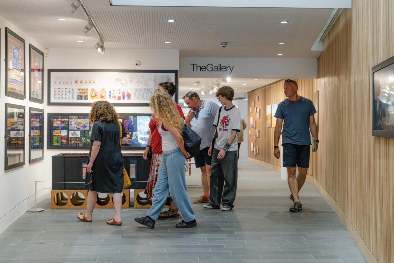 People browsing artwork in modern gallery space with white walls, track lighting, and "The Gallery" signage visible.