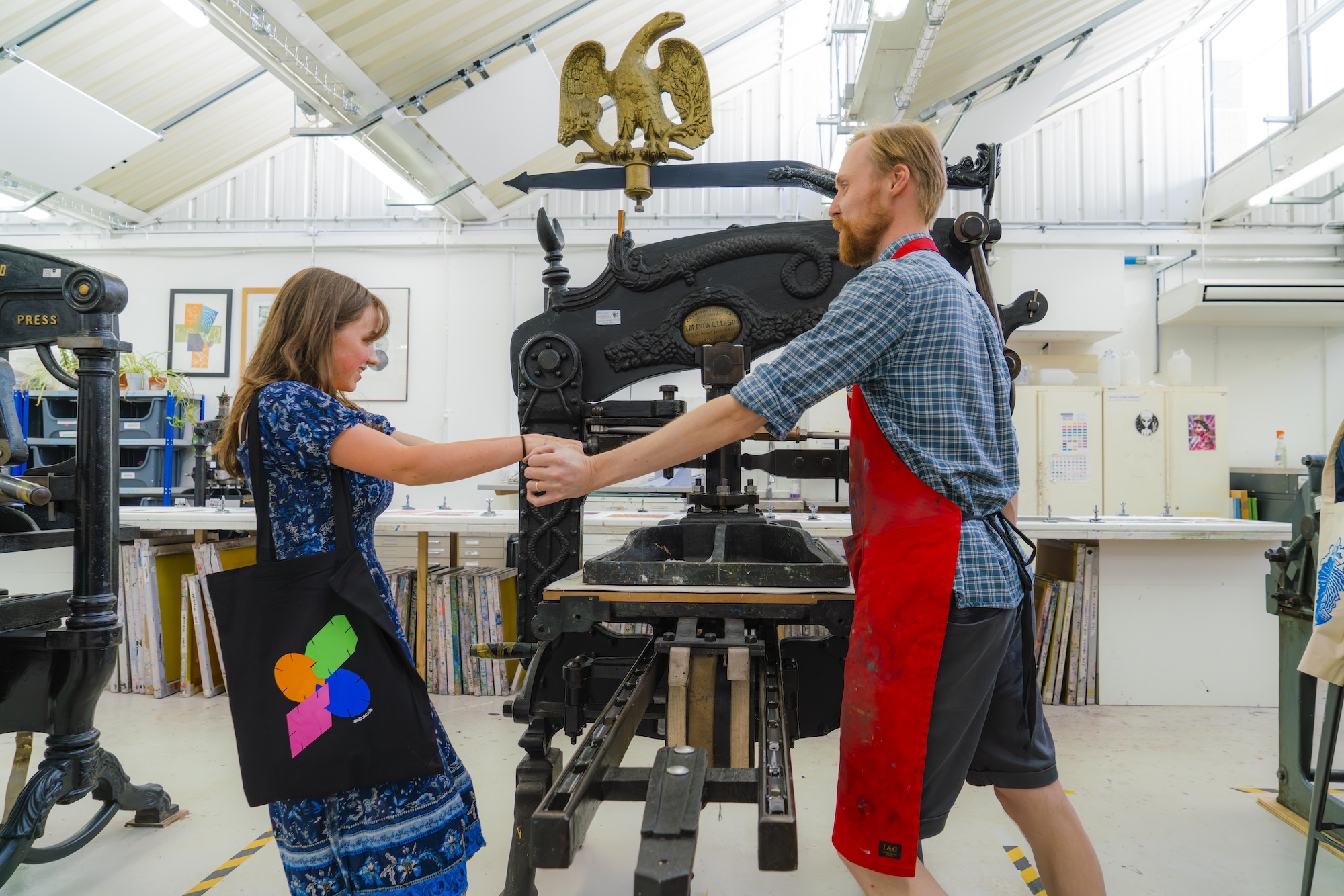 Two people operating a large black printing press in a workshop with white ceiling and walls. Woman in blue dress, man in checked shirt.
