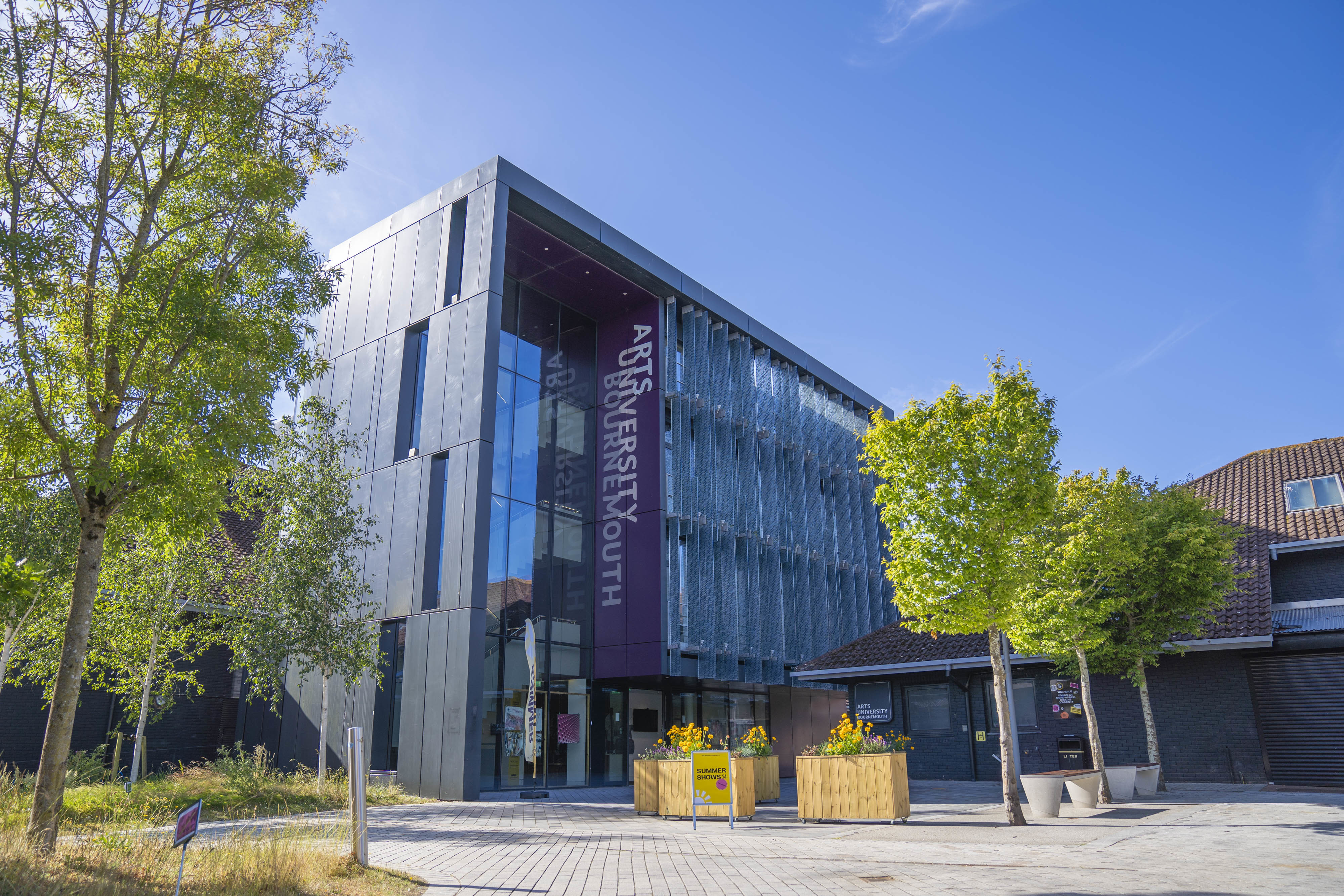 Exterior of the North Building on AUB Campus, with the words "Arts University Bournemouth" going vertically down a purple background on one side. A couple of trees line either side of the pavement in the foreground.