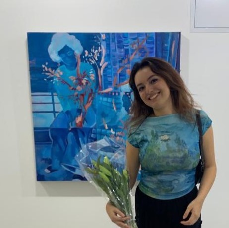 Liz Walker wearing a blue top and holding flowers in front of one of her blue, abstract paintings.