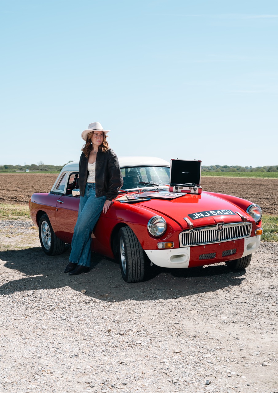 A woman wearing a hat and a black leather jacket leans against a red classic car with a white top. The car is parked on gravel with a countryside landscape featuring fields in the background under a clear blue sky.