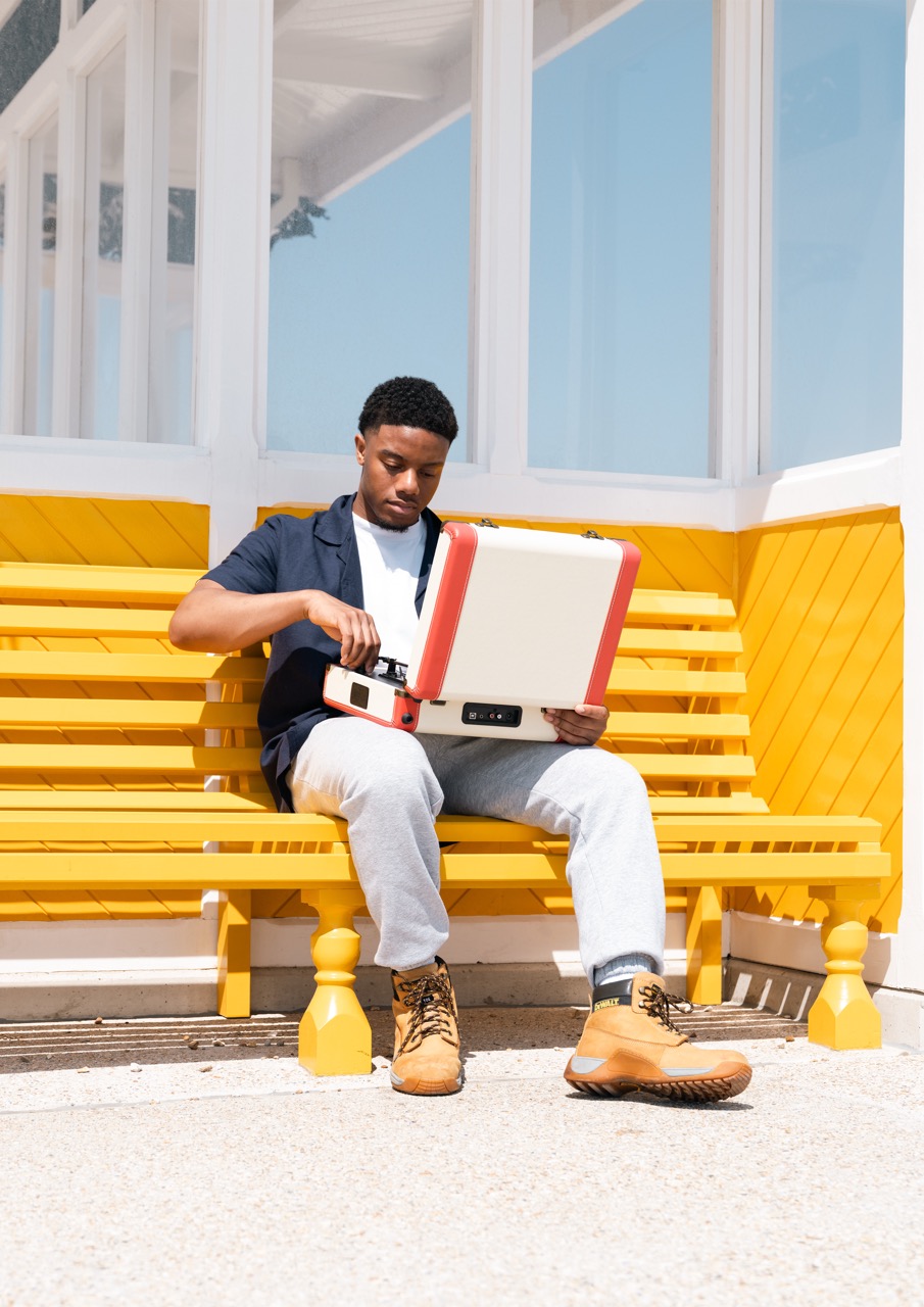 A young man sitting on a yellow bench in a vibrant setting, focused on opening a retro-style grey and red case. He is wearing a black shirt, grey sweatpants, and brown boots. The background features a bright, sunny atmosphere.
