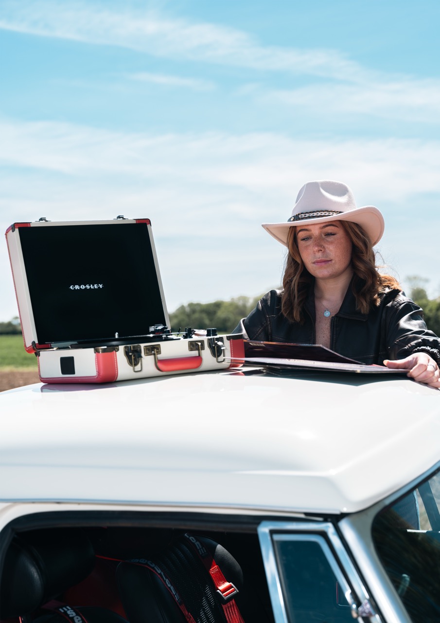 A woman wearing a white hat and black leather jacket stands beside a vintage car. A red and white portable record player is open on the car roof, with a scenic landscape visible in the background under a clear blue sky.
