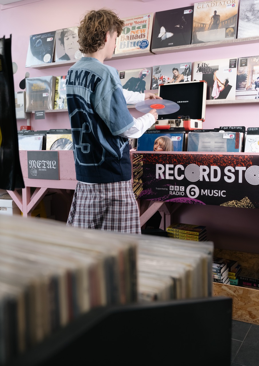 A young person with curly hair is holding a vinyl record in a record store. They are wearing a blue T-shirt with a number on the back and plaid shorts. The surrounding shelves display various vinyl records, while a sign reads "RECORD STORE."