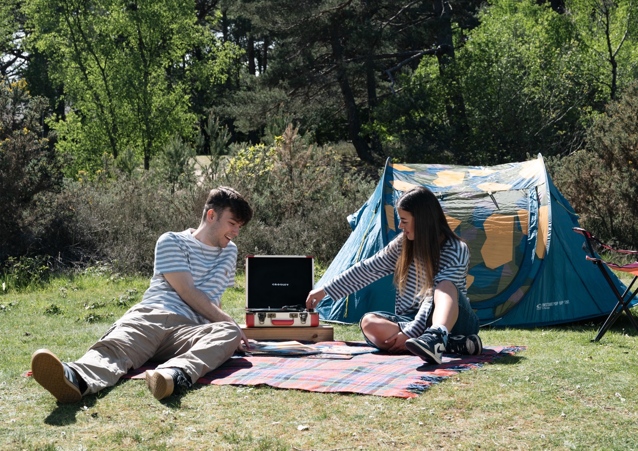 A young man in a striped shirt sits on a blanket, playing a record player while a young woman in a striped top leans over to adjust it. A blue and yellow tent is set up nearby in a lush green forest.