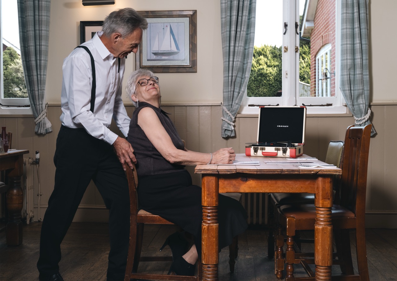 An older man in a white shirt and black trousers leans over a woman seated at a wooden table. The woman, wearing glasses and a black dress, faces away from the man. A record player sits on the table, with a window showing greenery in the background.