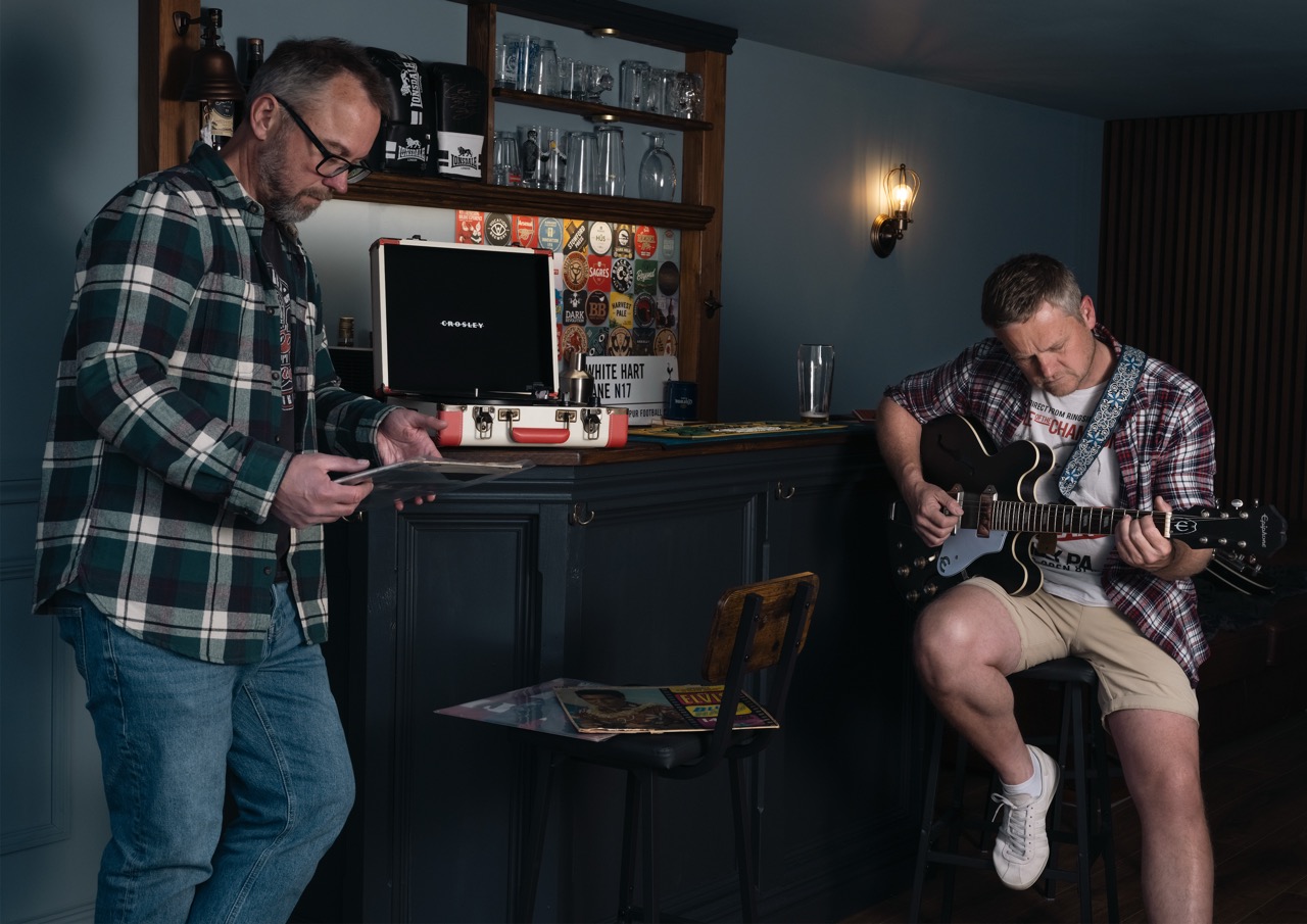 A man in a plaid shirt and glasses stands beside a bar, using a smartphone. Another man in a checked shirt and shorts plays an acoustic guitar while seated on a stool. The background features a bar with various drinks and illuminated decor.