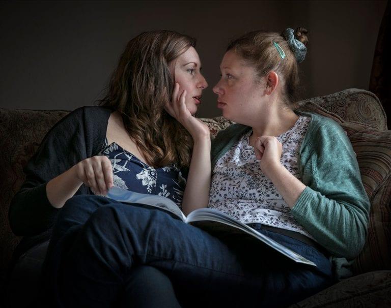 Two women on a sofa reading a book, close to embrace.