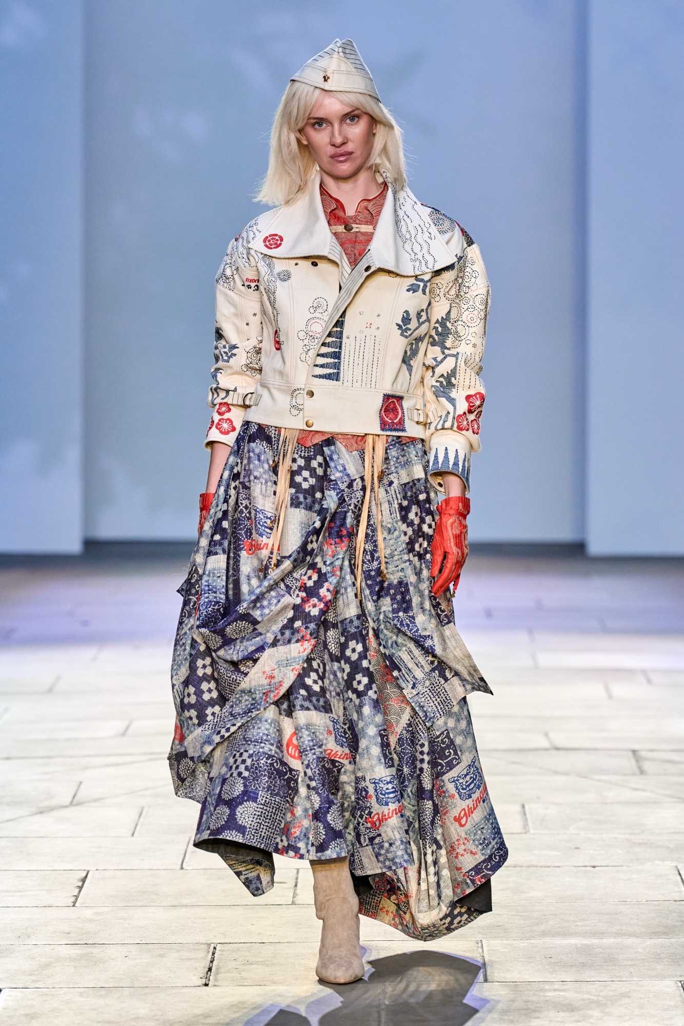 A blonde model wearing a patterned jacket, skirt and hat walks down a runway in a fashion show.