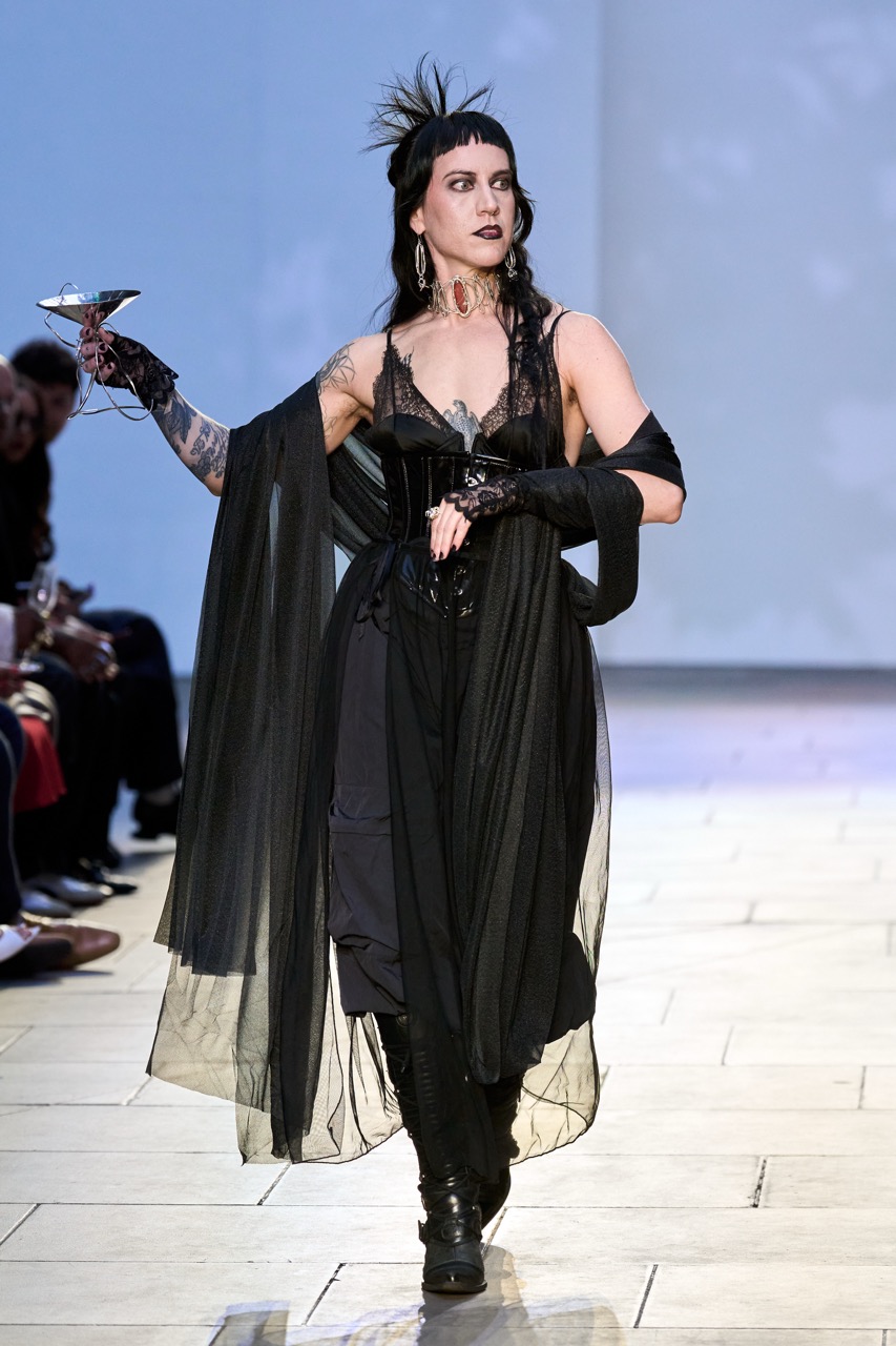 A model in a black sheer corset dress and draped cape walks a runway, holding a cocktail glass and wearing a feathered headpiece.