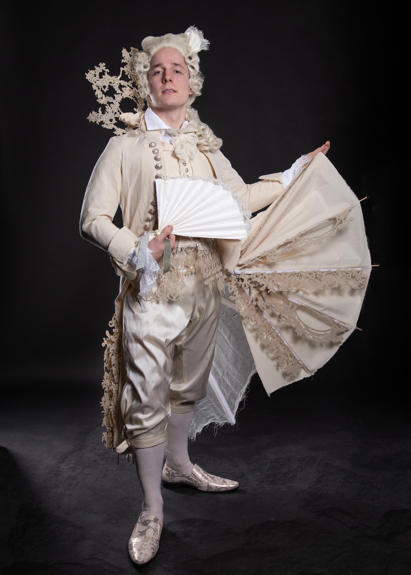 Model in an 18th century-style costume as Figaro, facing the camera with a fan in one hand and one panel of his tailcoat, spread out like a wing, in the other hand.