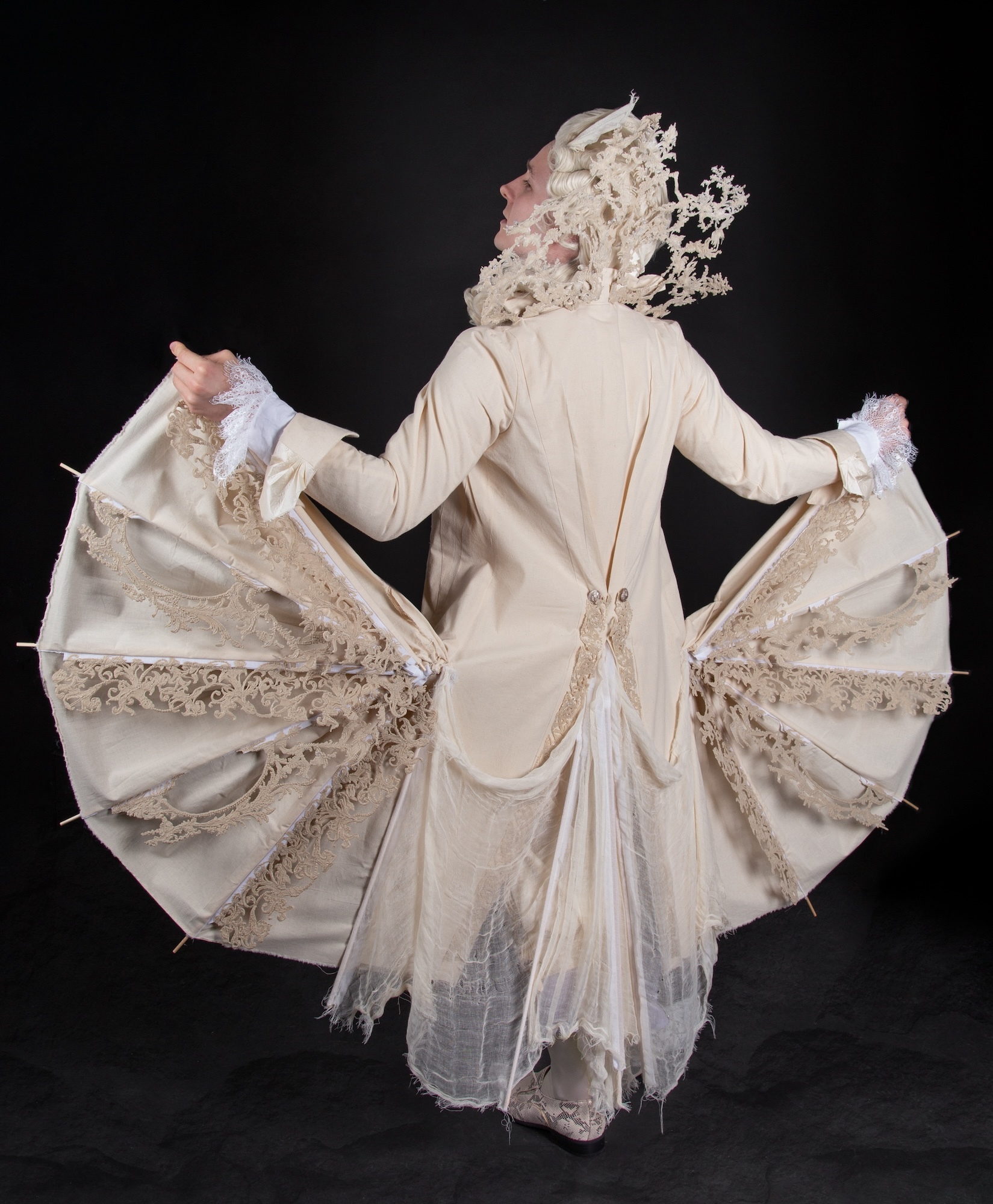 Model in a cream 18th century-style tailcoat as Figaro, his back to the camera with the panels of his coat spread out like wings.
