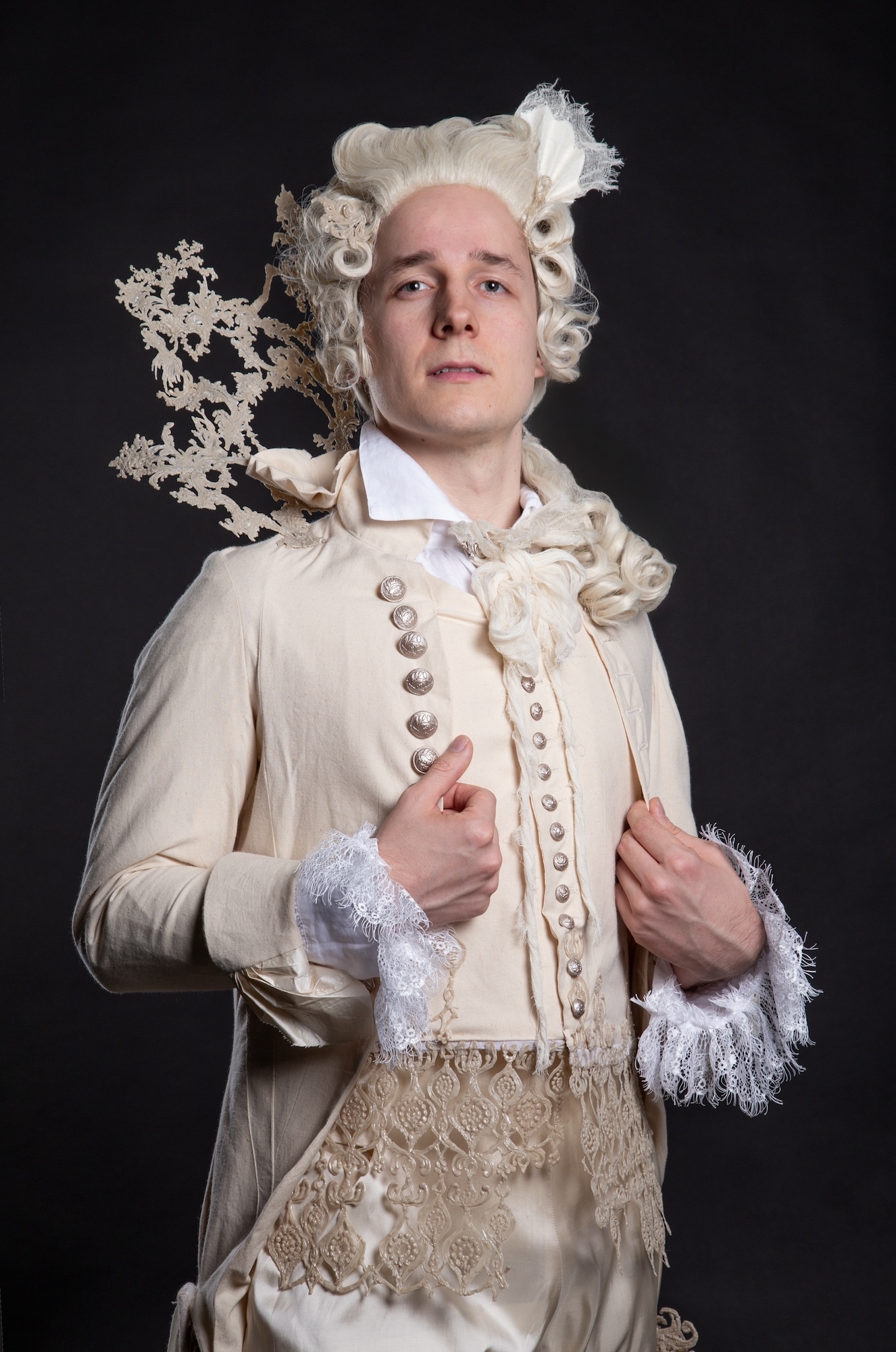 Model wearing a cream-coloured 18th century costume and powdered wig, posing with his hands on his lapels.