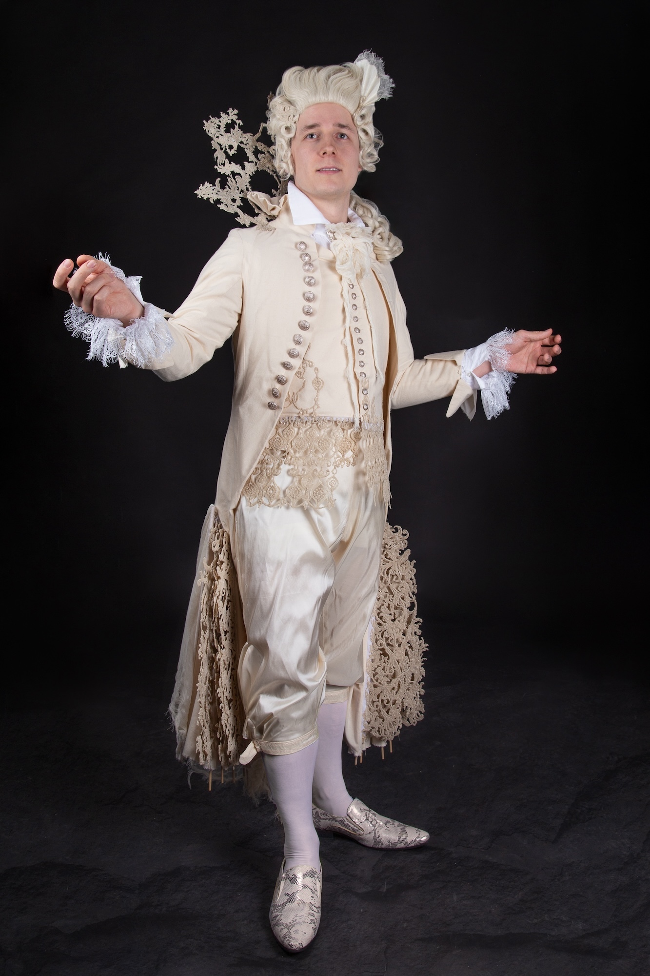 A model in an 18th century-style costume as Figaro faces the camera with raised arms.