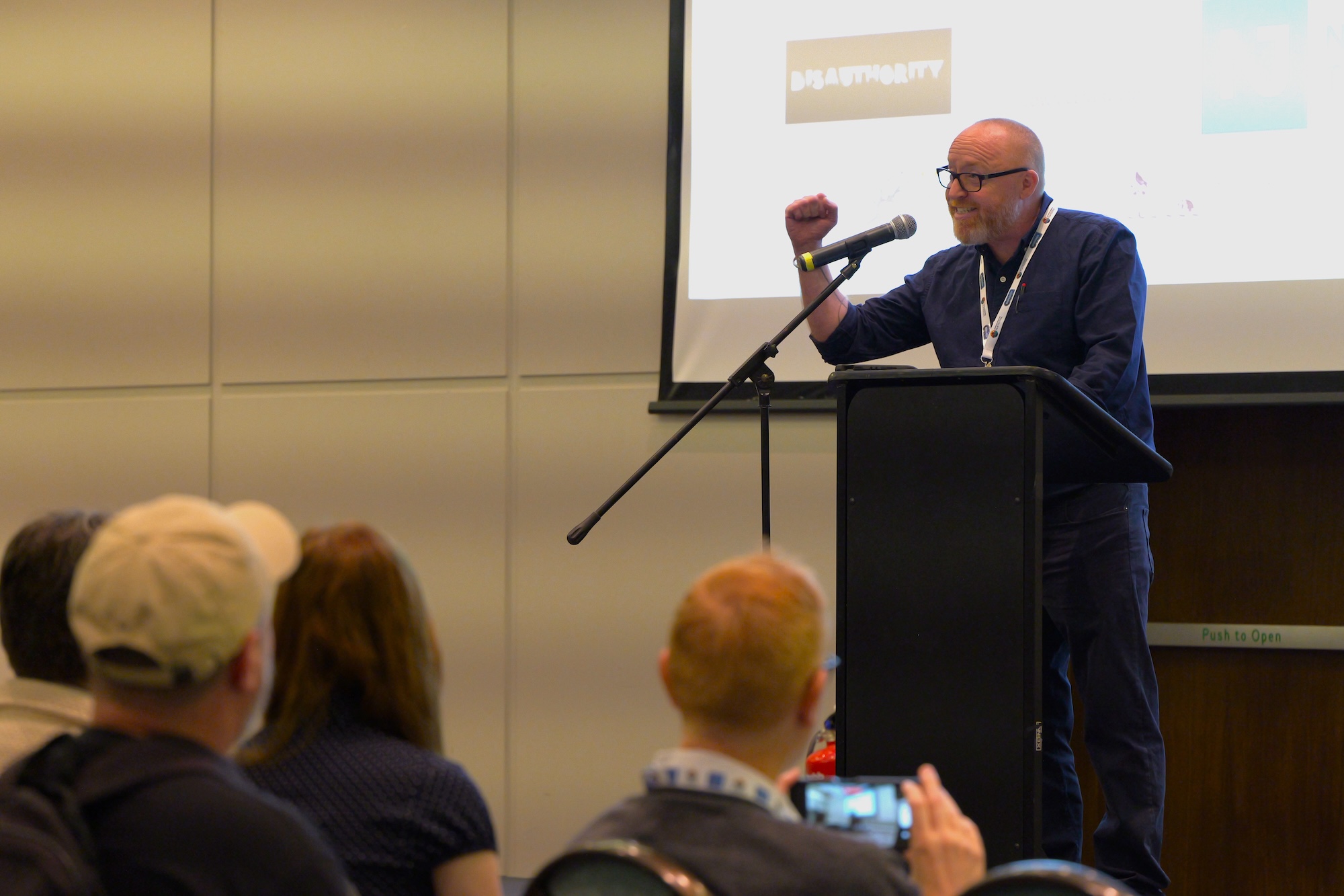 A person speaking at a podium with a microphone in a conference room, with seated audience members in the foreground and a presentation screen behind him.