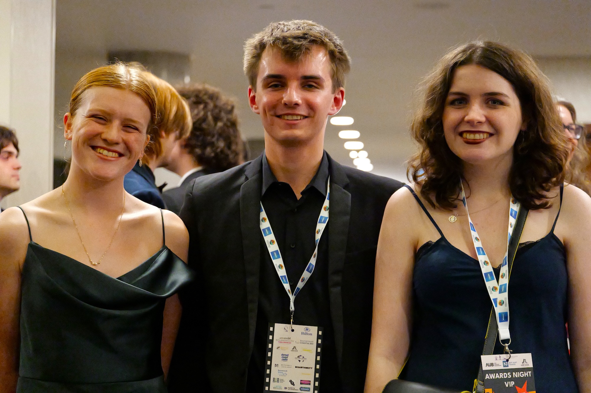 Three young adults in eveningwear and lanyards pose indoors at an event, with other attendees and corridor lights in the background.