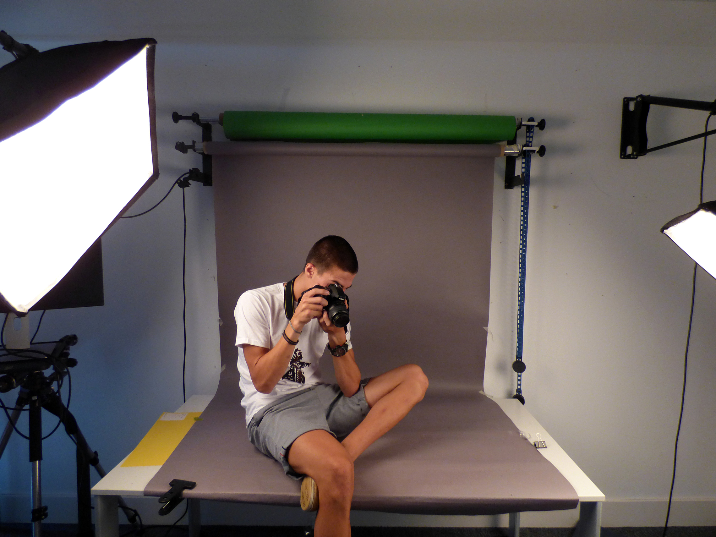 A student is sitting in front of a green screen backdrop pointing a camera at the ground.