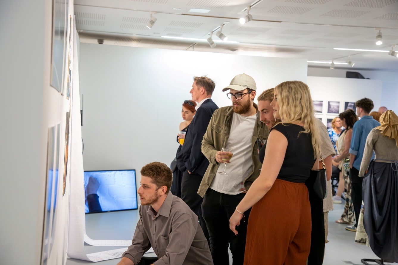 Gallery visitors viewing wall-mounted displays and digital screen. White walls, bright lighting, people in casual clothing gathered around exhibits.
