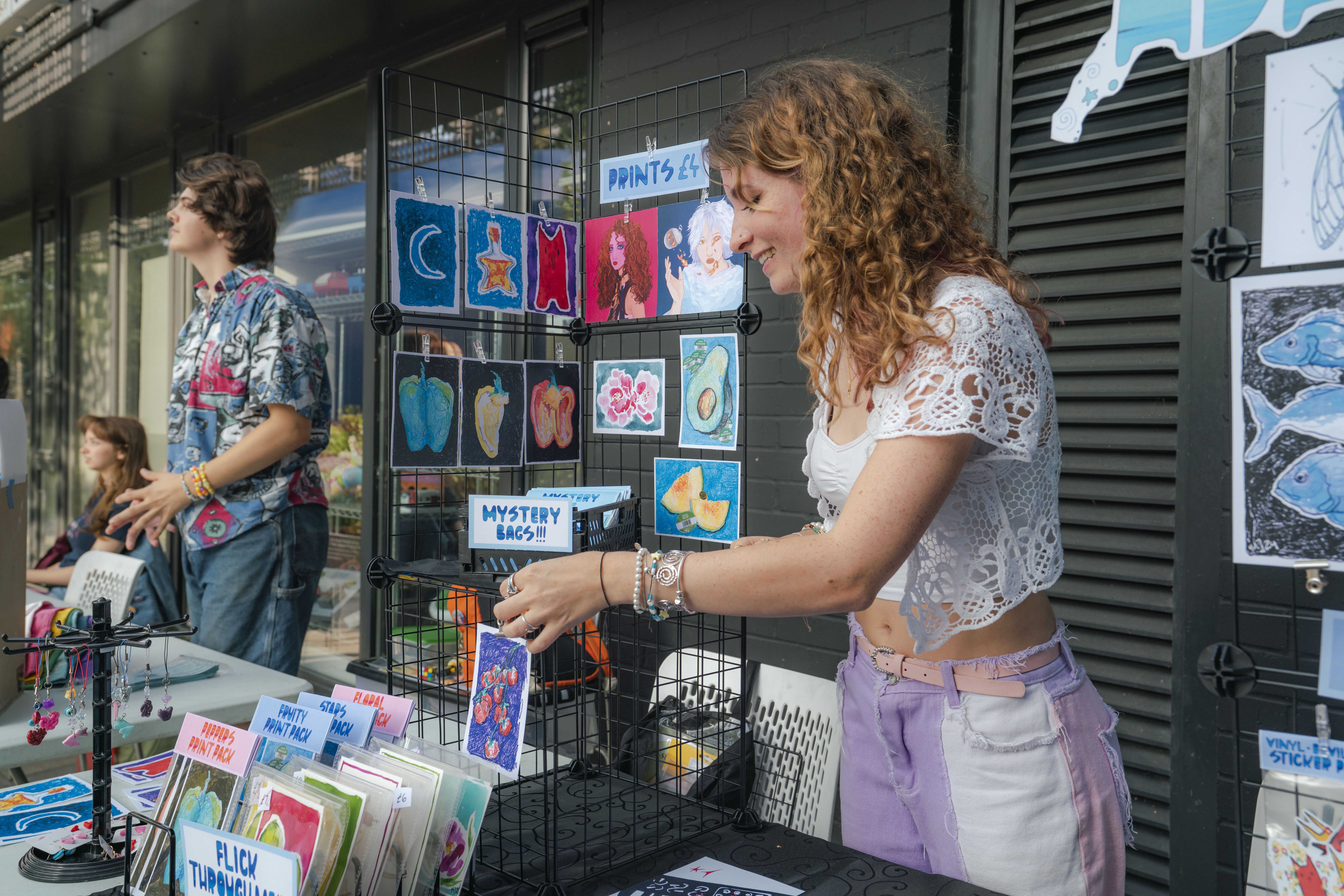 Student with curly hair and white top stood in front of stand with colourful, painted illustrations, smiling and talking.