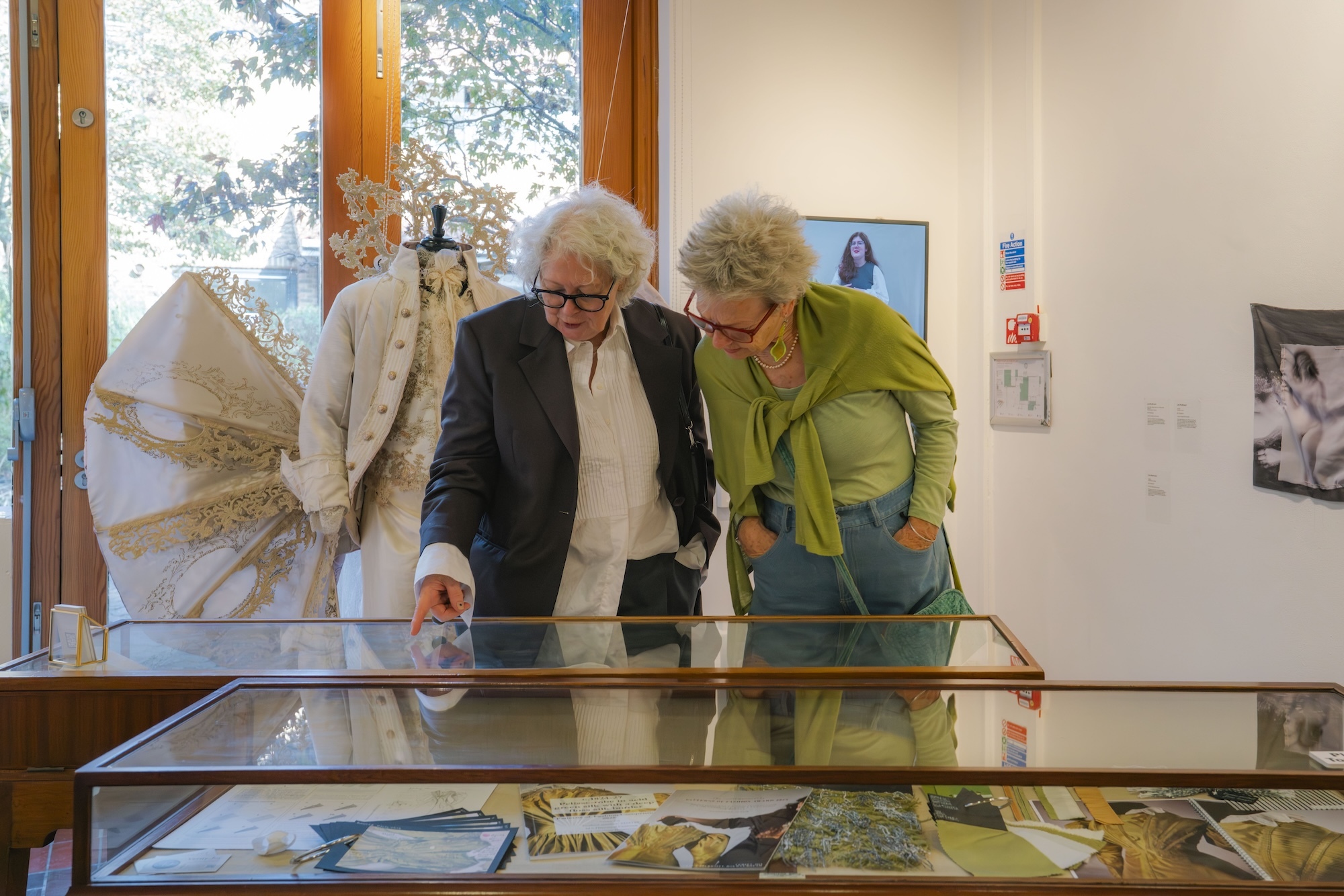 Two elderly people examining items in glass display case at museum or gallery with white walls and large windows.