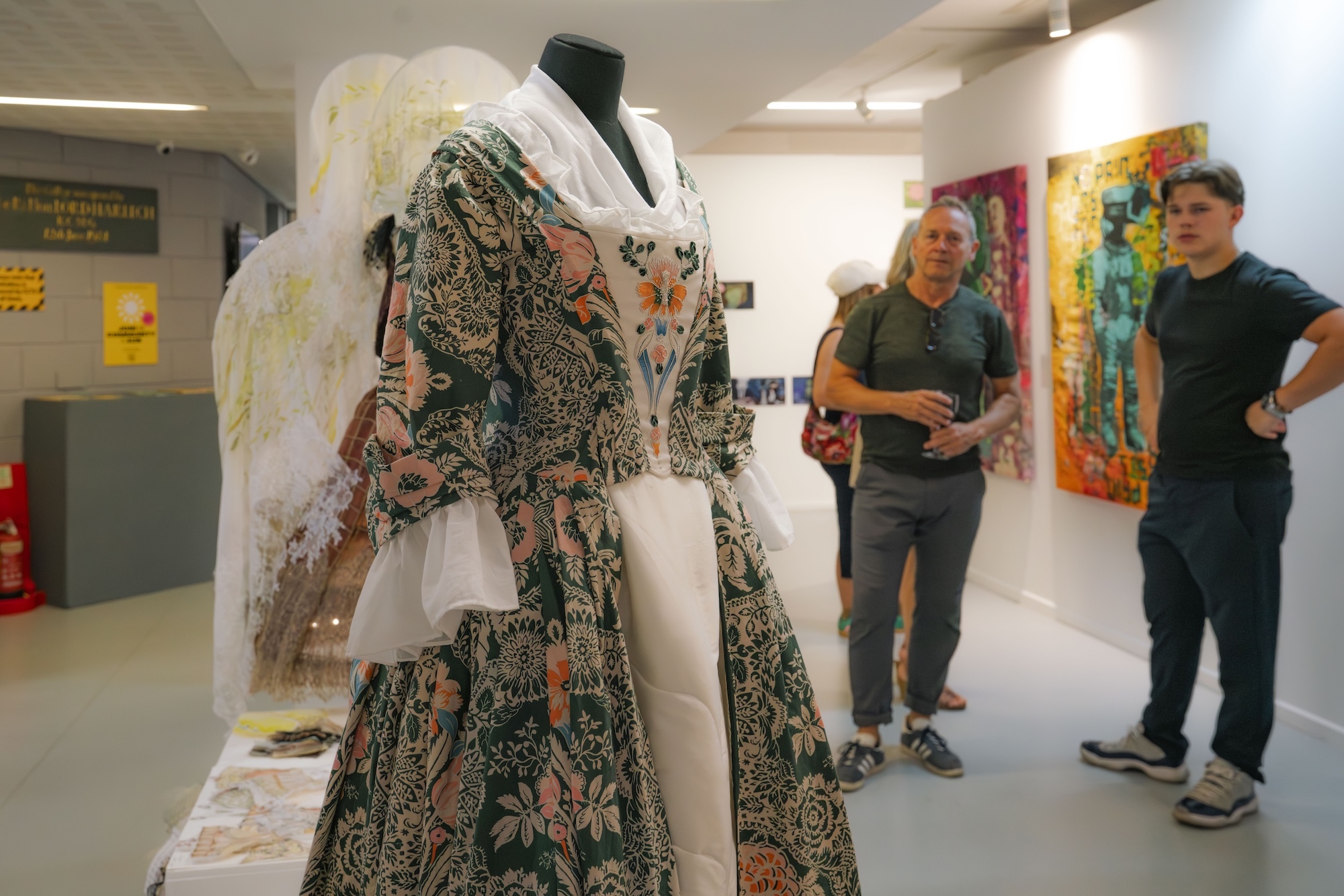 White elephant sculpture covered in patterned fabric and decorations displayed in gallery with visitors viewing colourful artwork on walls.