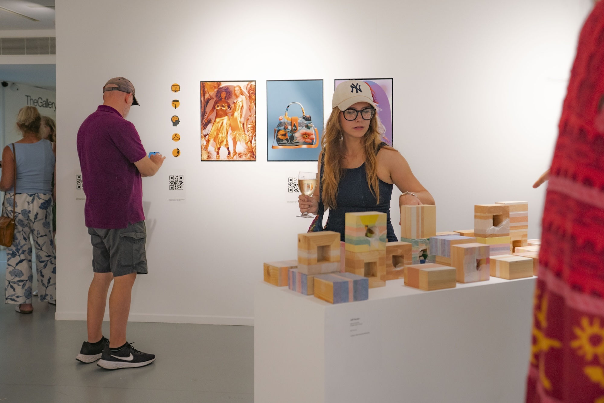 Gallery interior with white walls displaying colourful artwork. Woman with long hair and glasses stands behind white table with wooden blocks. Visitors in purple and red clothing view exhibits.