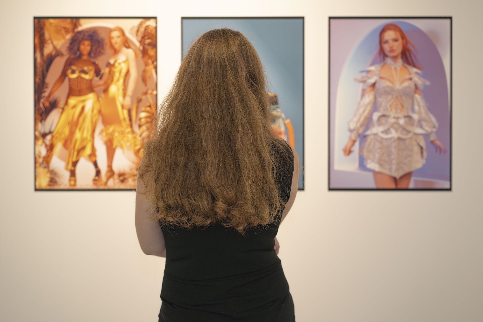 Person in black top viewing three framed artworks on white gallery wall.
