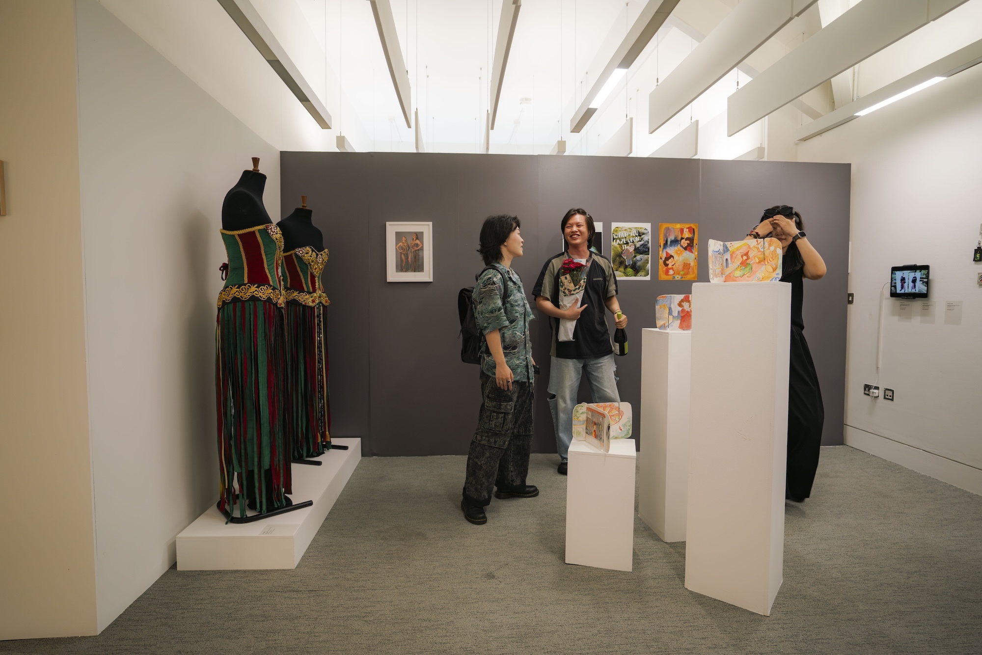 Gallery interior with white exposed beam ceiling, grey feature wall displaying framed artwork, white display pedestals, and three visitors examining exhibits.