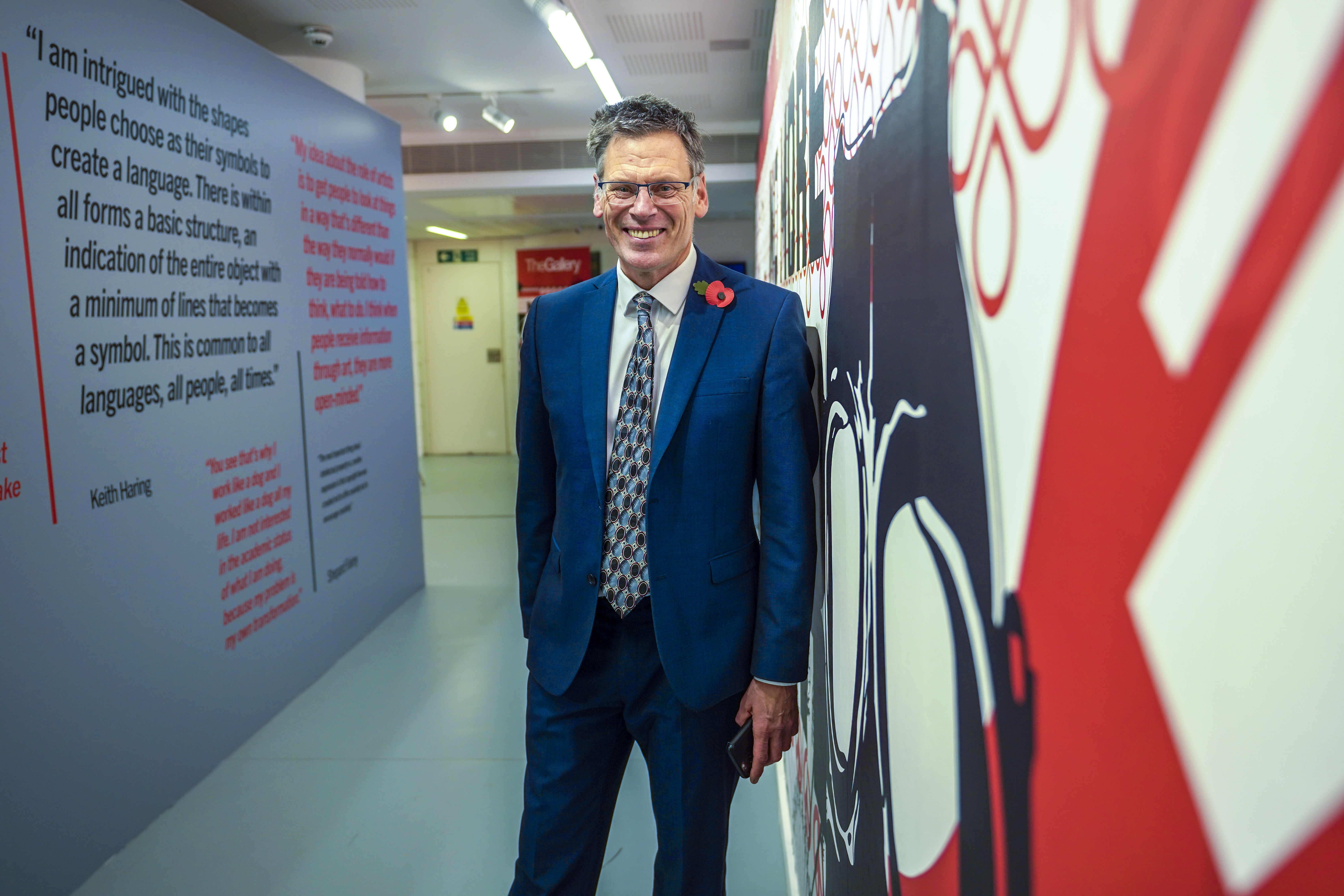 Photo of Professor Paul Gough, standing next to a large street artwork printed on a wall. Blocks of text are printed on the wall visible on the left-hand side.